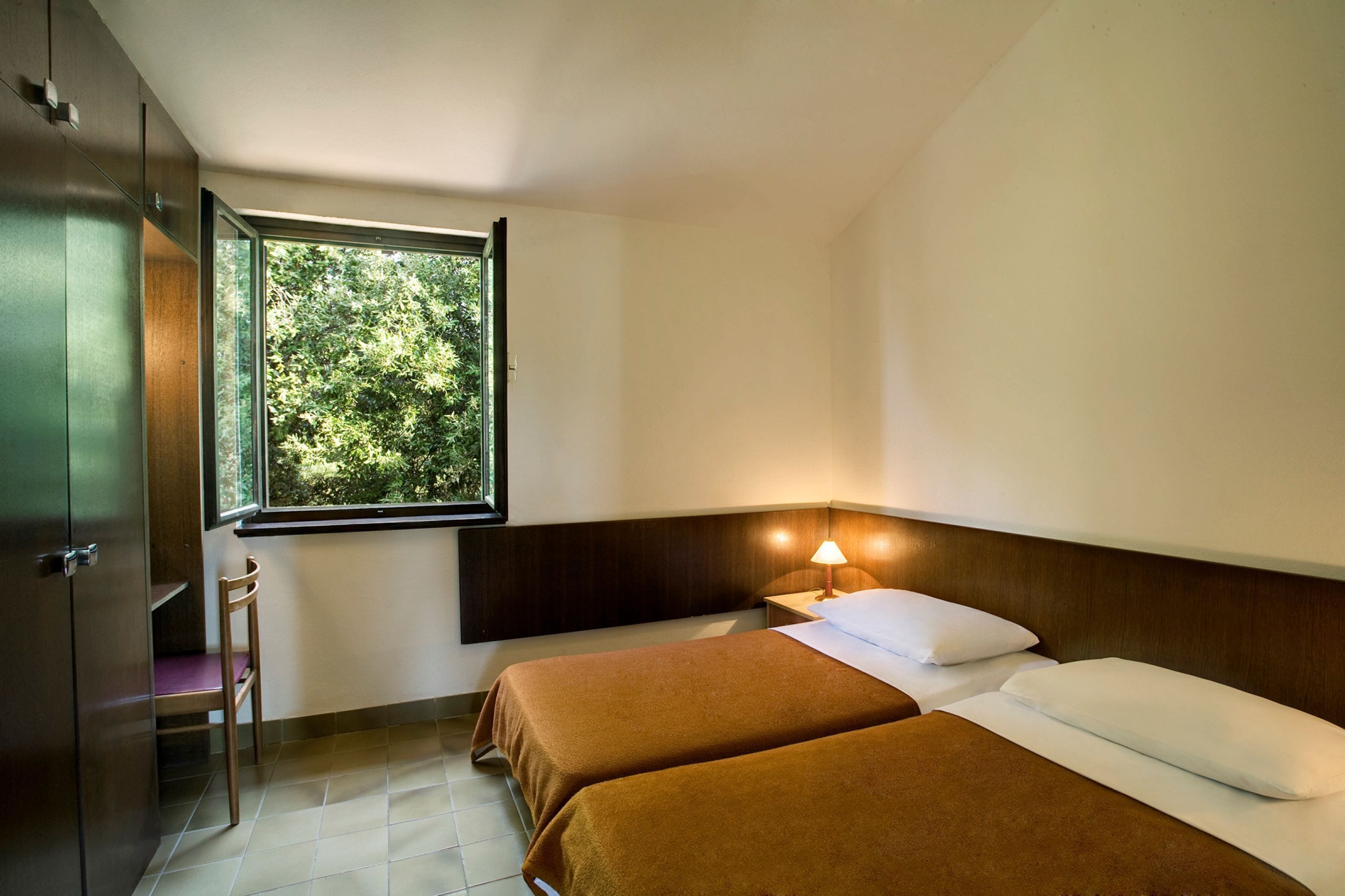A hotel room with two beds, a window with a view of greenery, a chair, and a nightstand with a lamp.