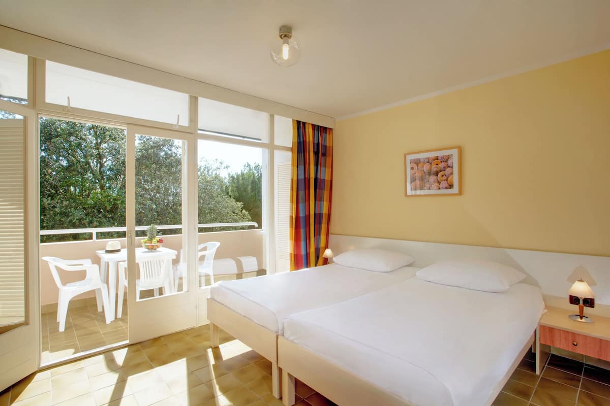 A hotel room with a bed, a bedside table with a lamp, a window with curtains, a balcony with chairs and a table, and a painting on the wall.
