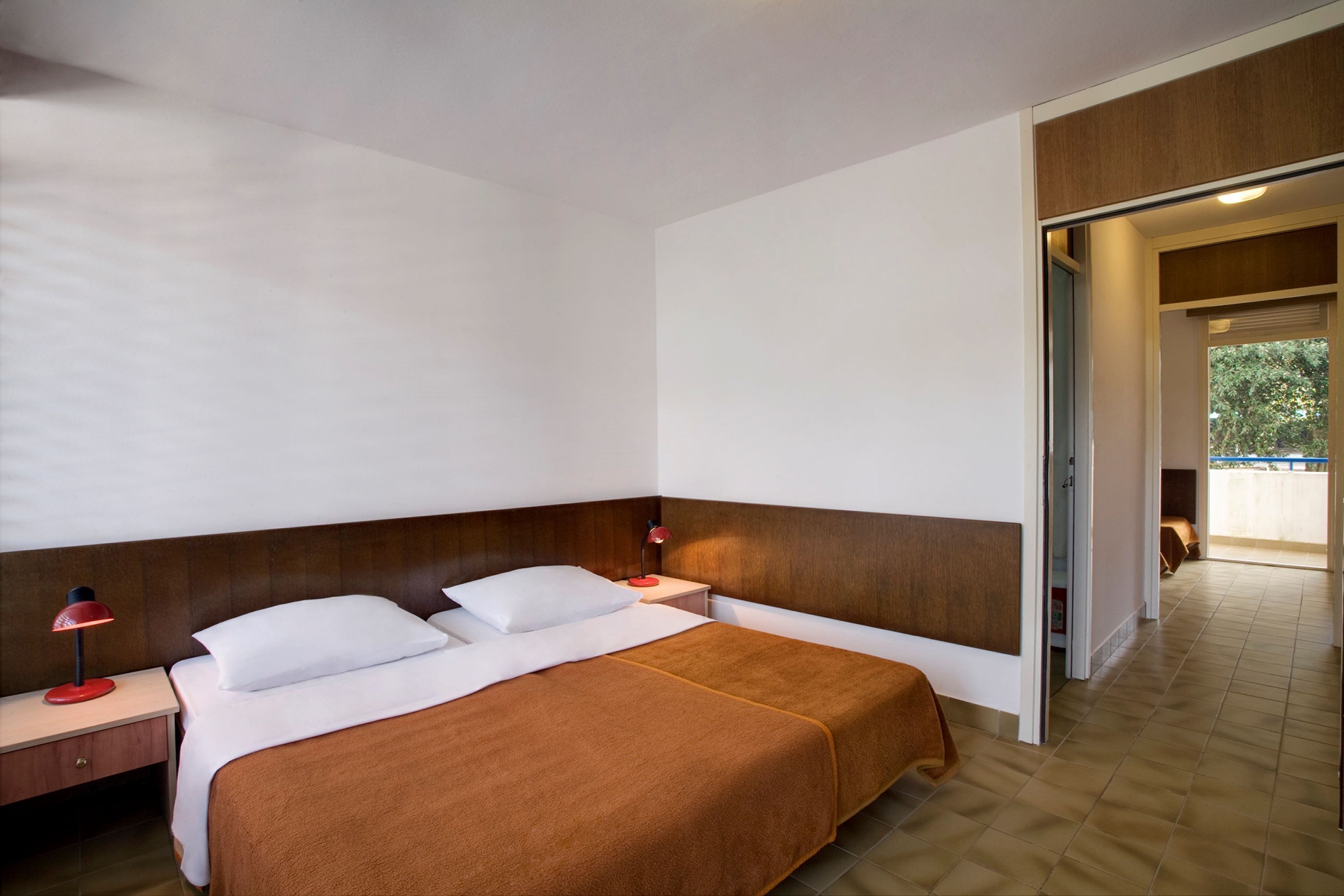 A hotel room with a bed, a nightstand, a red lamp, and a wooden headboard.