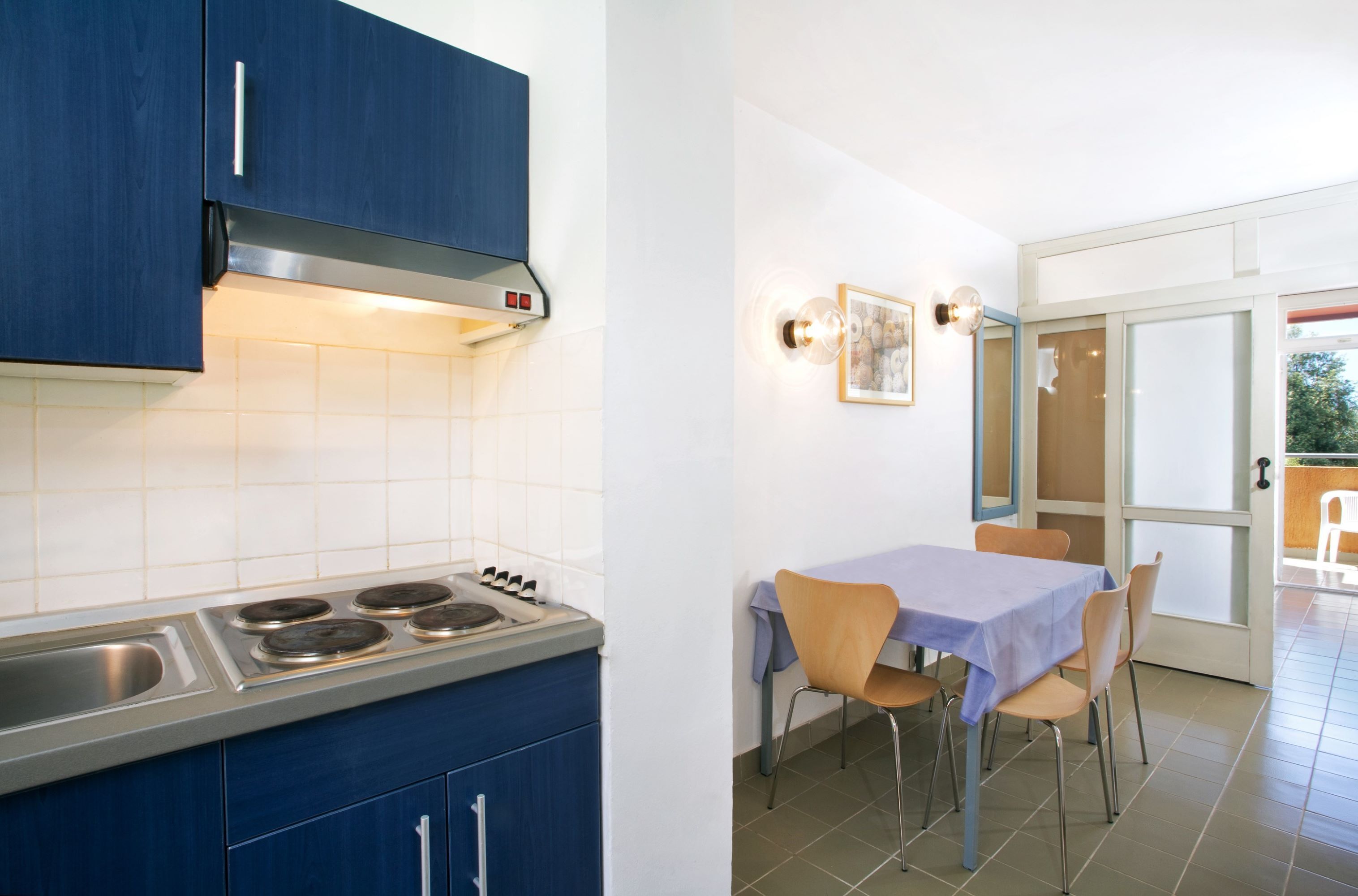 Eine Küche mit Herd, Spüle und blauen Schränken. Ein Esstisch mit Stühlen und ein Fenster mit Blick ins Freie.