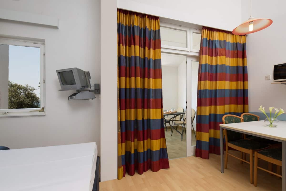A room with a bed, a desk with a chair, a television on a shelf, and a window with curtains.