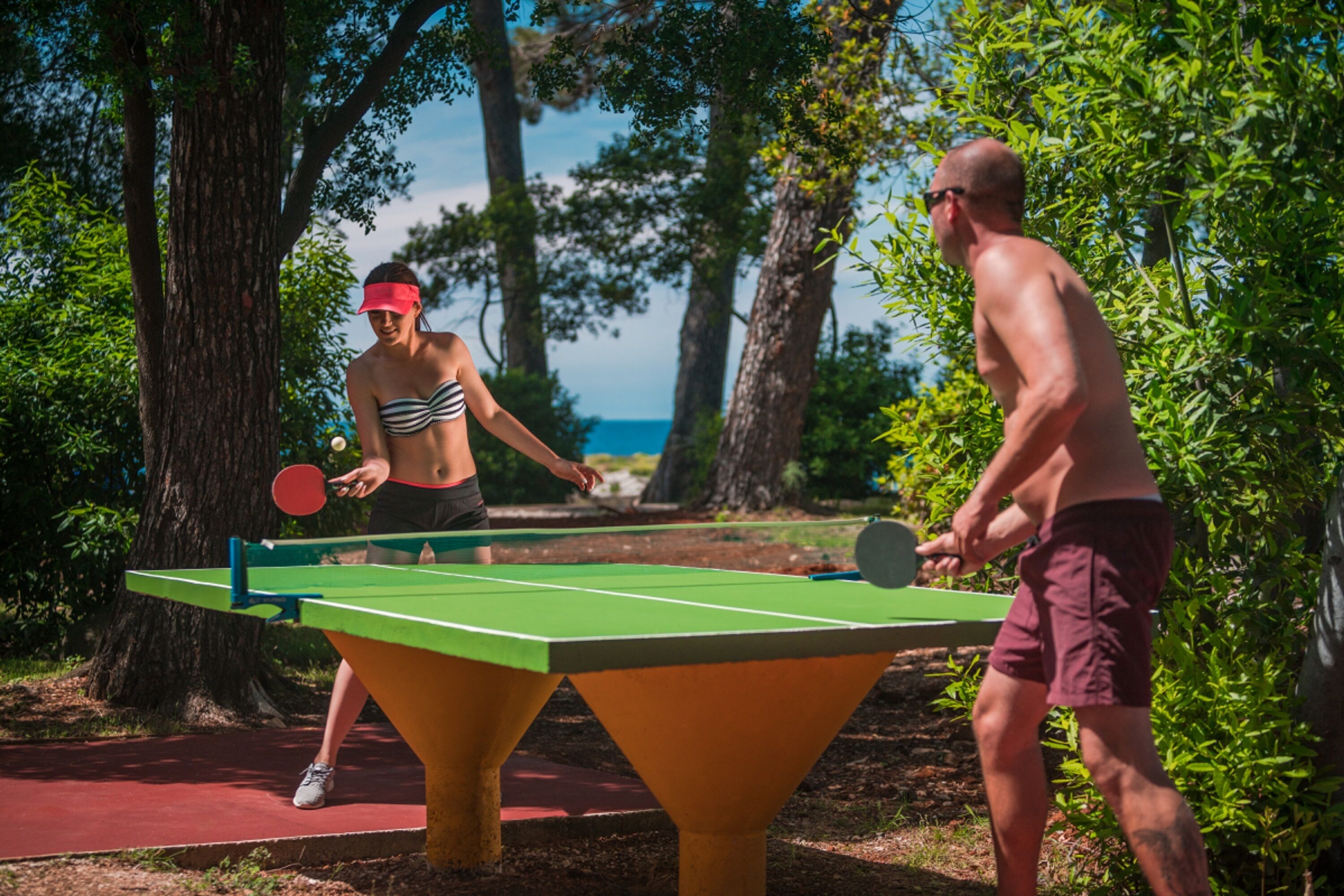 Due persone giocano a ping pong all'aperto, con un tavolo verde e un pavimento rosso, circondati da alberi e un cielo sereno.