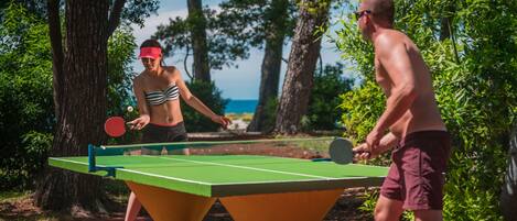 Two people playing table tennis outdoors with a green table and red floor, surrounded by trees and a clear sky.
