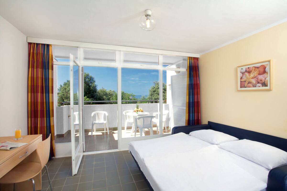 A hotel room with a bed, a desk with a chair, a table with chairs, and a balcony with a table and chairs.