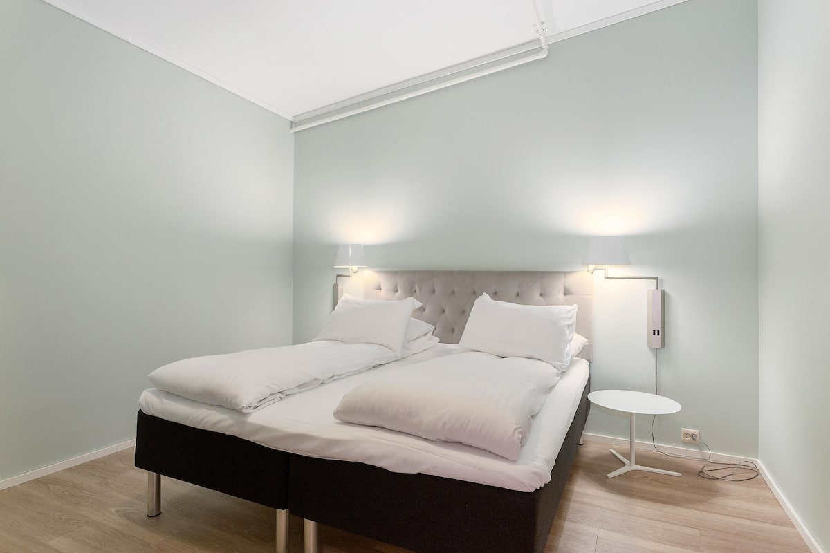 Una cama bien hecha con sábanas blancas y cabecera negra, una mesita redonda con lámpara, y un enchufe de pared.