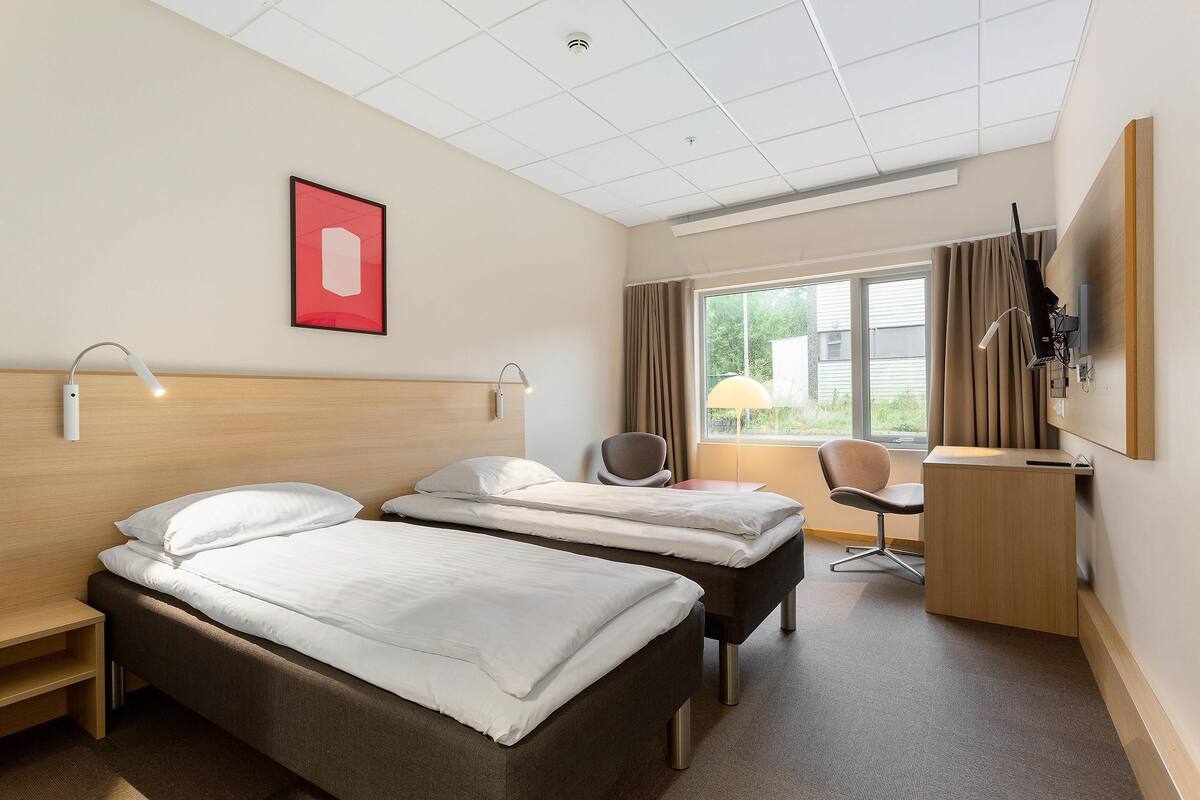 Habitación de hotel con dos camas, un escritorio, una silla, televisión y ventana con cortinas.