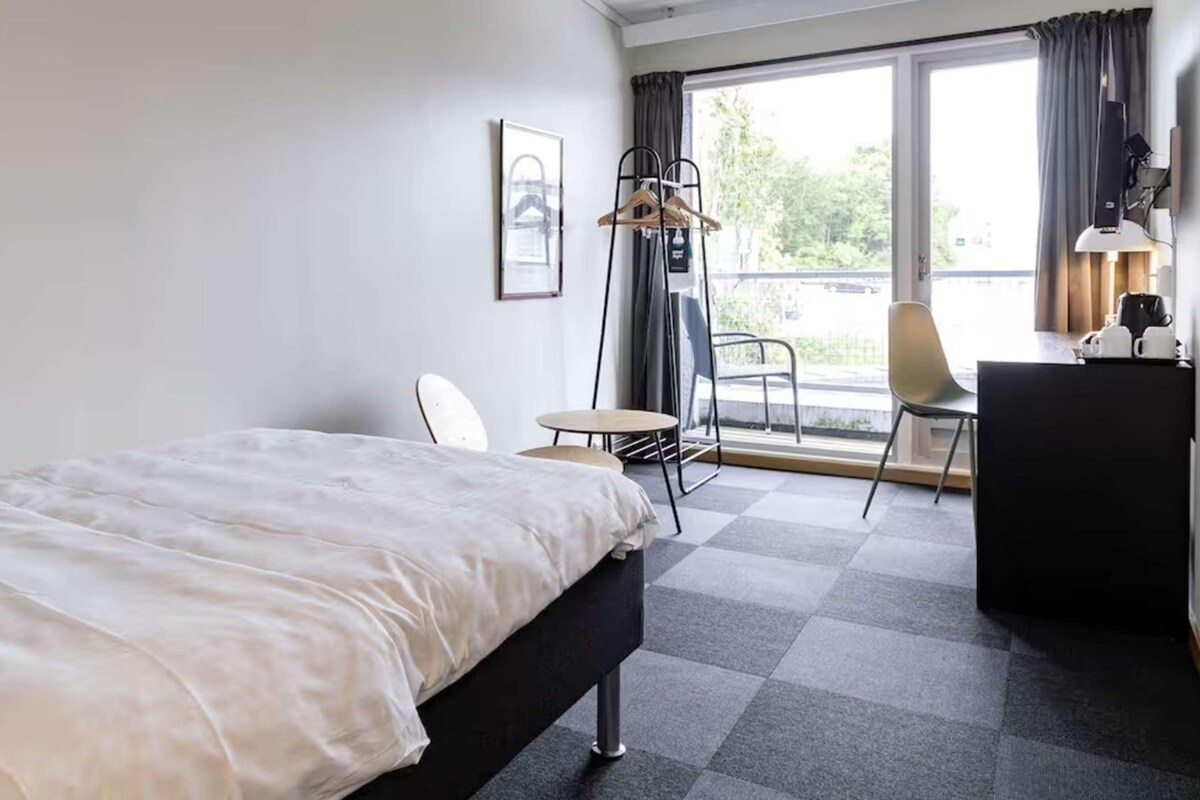 Quarto de hotel com cama, escrivaninha, cadeira, mesa pequena e vista para o exterior.