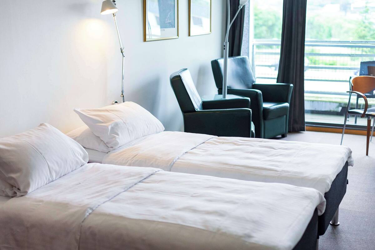 Habitación de hotel con dos camas, un escritorio, una silla y una ventana con vista a zonas verdes.