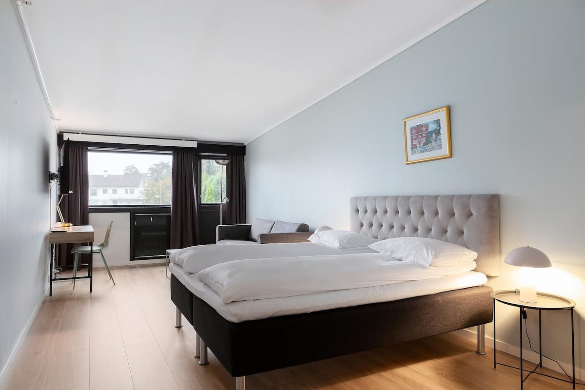 Un dormitorio con cama, escritorio, silla, mesita de noche con lámpara y ventana con cortinas.