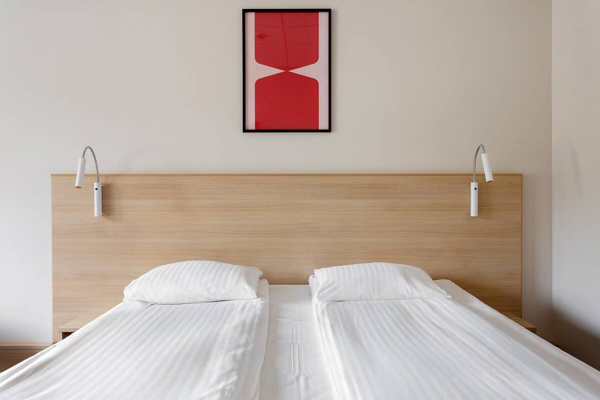 Una cama bien hecha con sábanas blancas, dos lámparas de noche, un cabecero de madera y una obra de arte abstracta enmarcada en la pared.