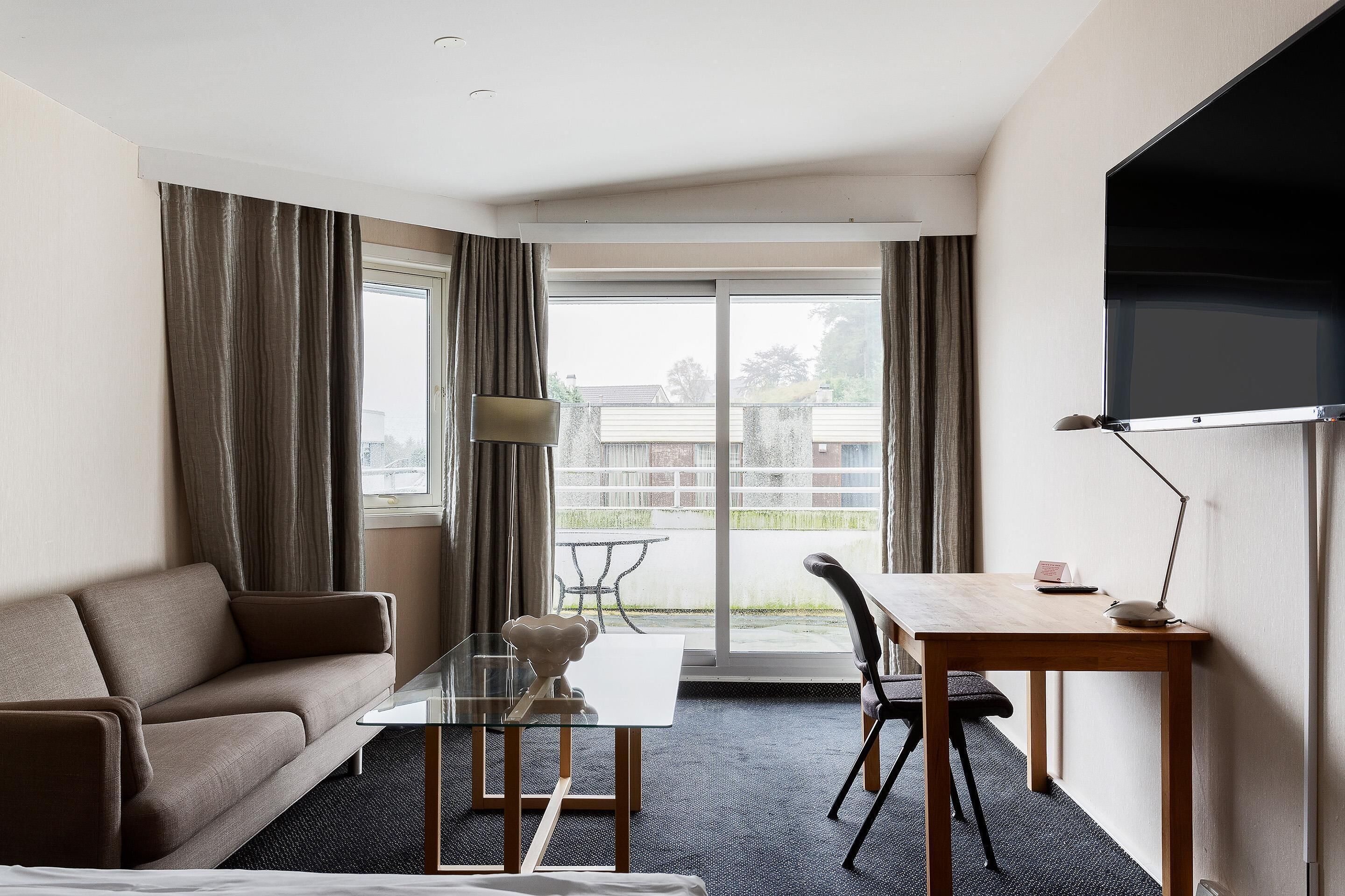 A hotel room with a sofa, a glass-top table, a chair, a desk, a TV, and a balcony with a table and chairs.