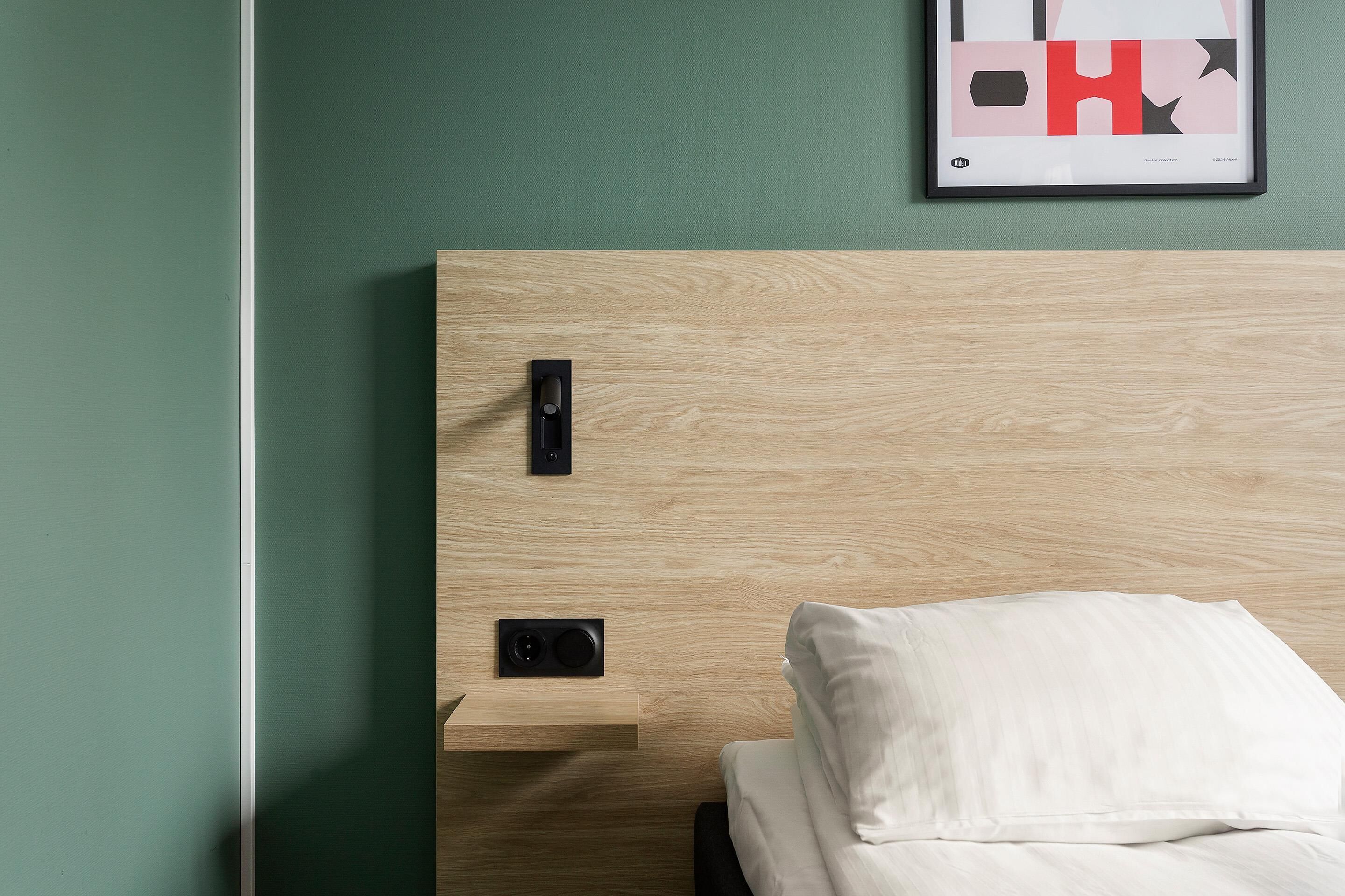 A hotel room with a wooden headboard, a bedside table with a lamp, and a framed abstract artwork on the wall.