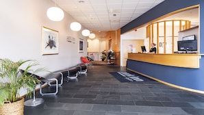 Zona de recepción con mostrador de madera, una recepcionista y un área de espera con sillas y un mostrador.