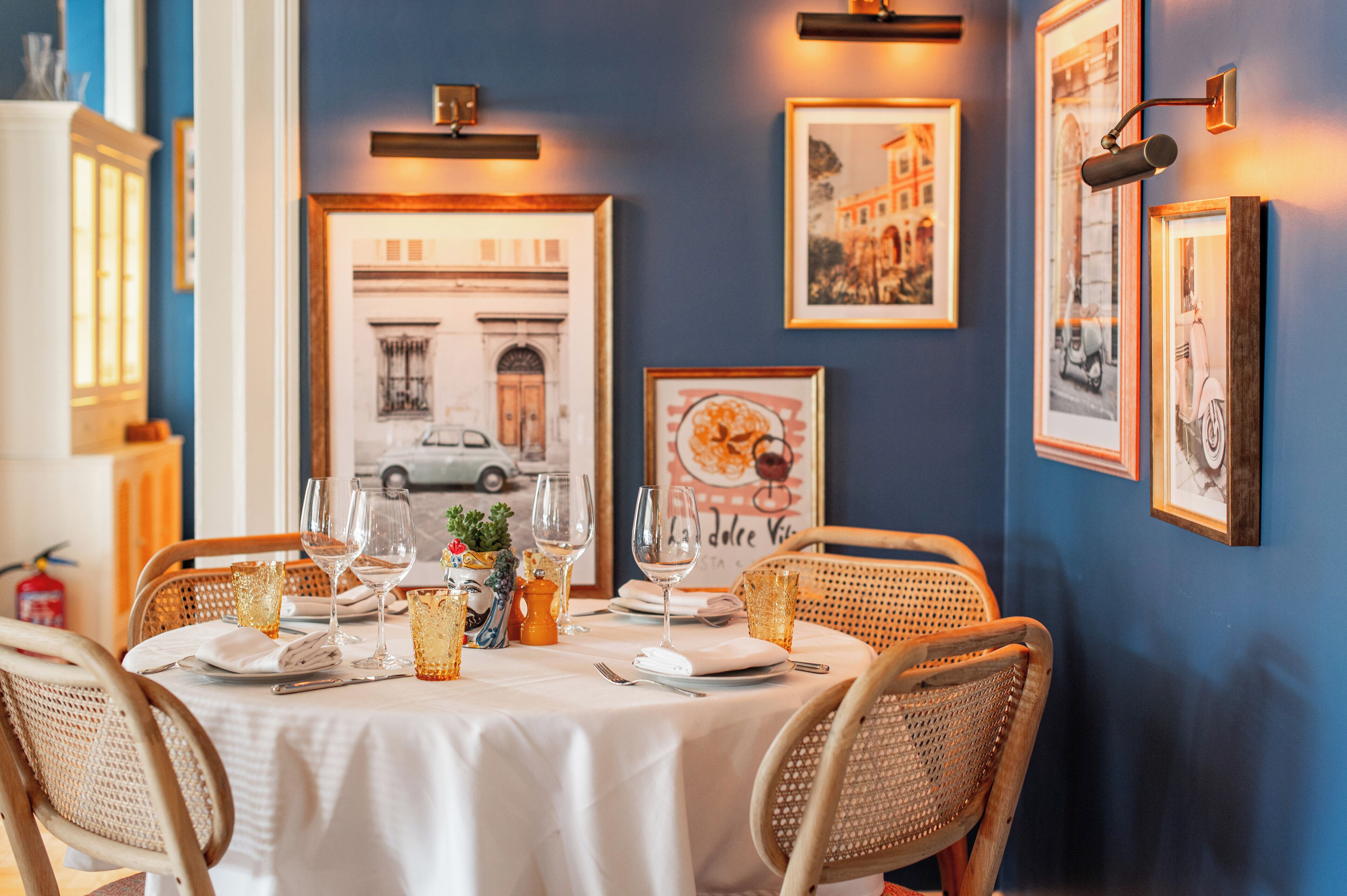 Un coin repas avec une table dressée pour deux personnes, des tableaux encadrés au mur et une vue sur un bâtiment par la fenêtre.