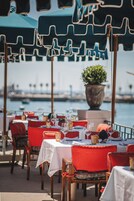 Un área de comedor al aire libre con mesas preparadas para dos, sillas rojas y sombrillas verdes que dan sombra.