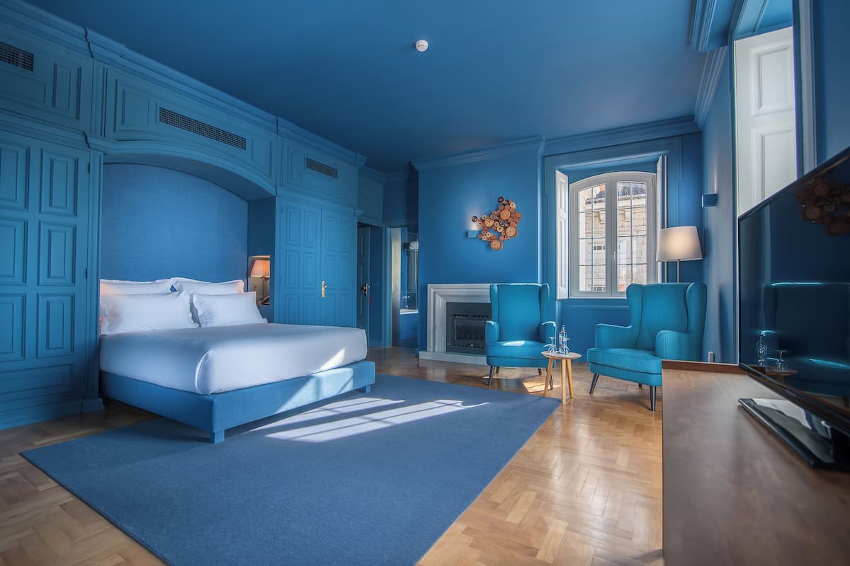 Un dormitorio con una cama grande, dos sillones azules, un aparador de madera y una chimenea.