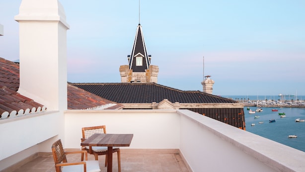 Un balcón con mesa y sillas, con vista a un puerto deportivo con barcos y una edificación distante con techo puntiagudo.
