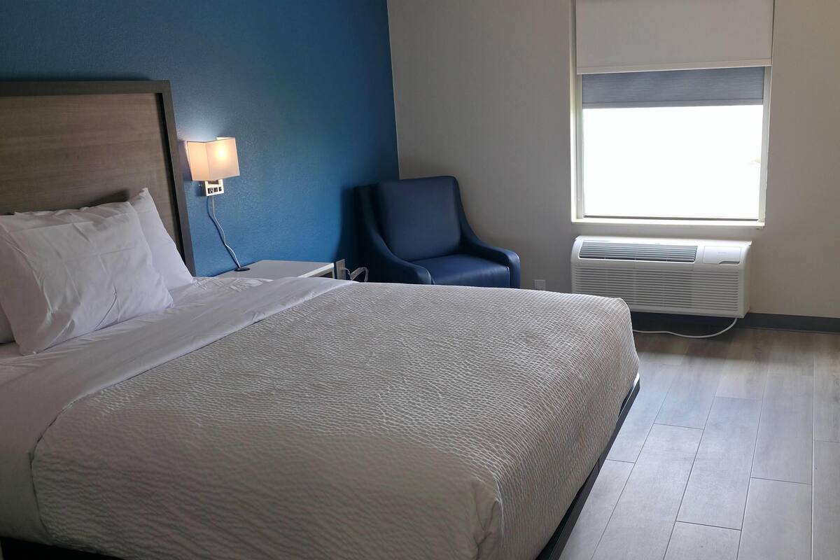 A hotel room with a bed, a chair, a lamp, and an air conditioning unit.