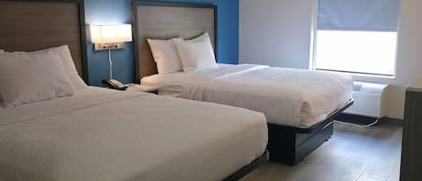 A hotel room with two beds, a nightstand with a phone, a wall-mounted lamp, and a window with a blind.