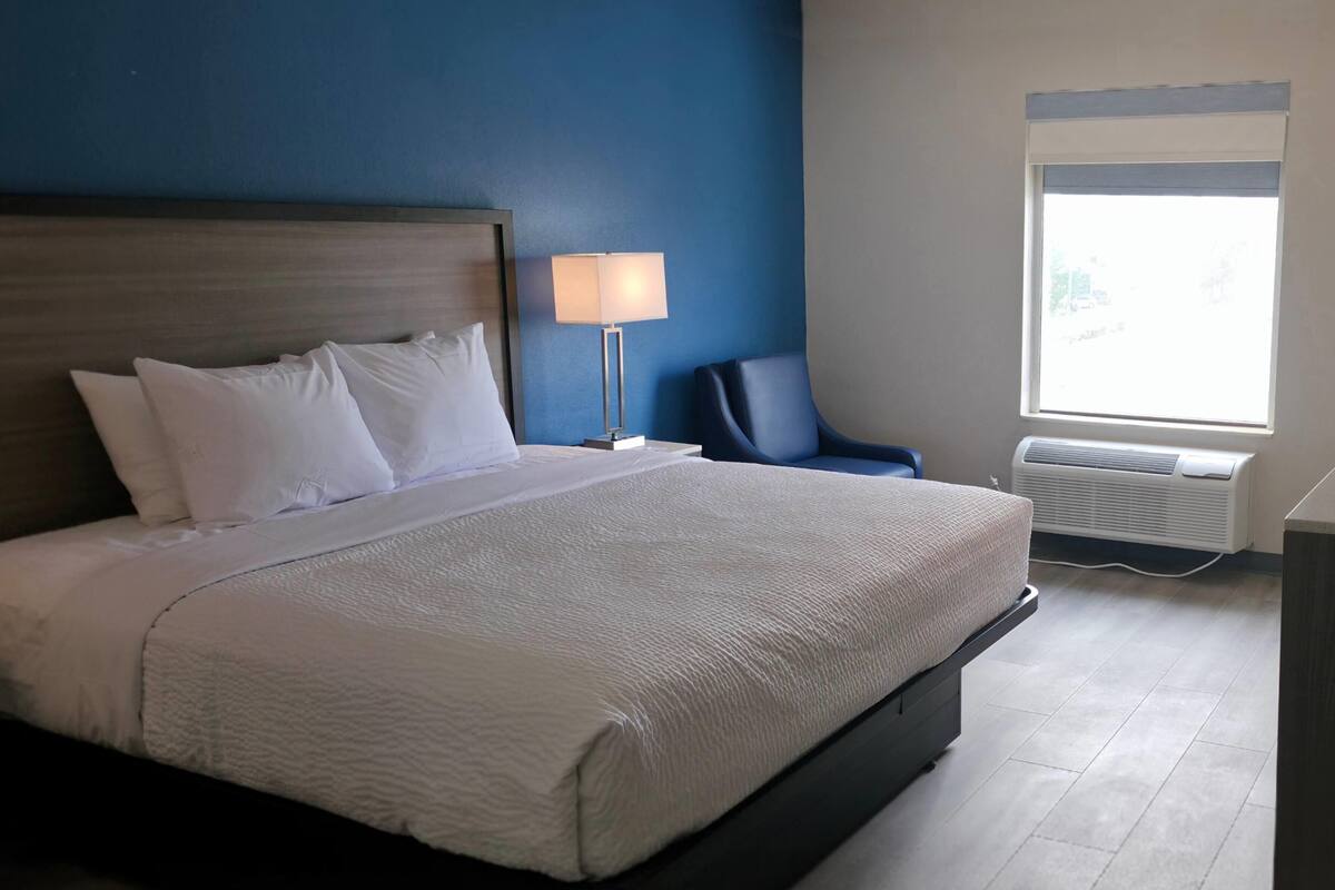 A neatly made bed with white linens, a bedside lamp, a chair, and a window with an air conditioning unit.