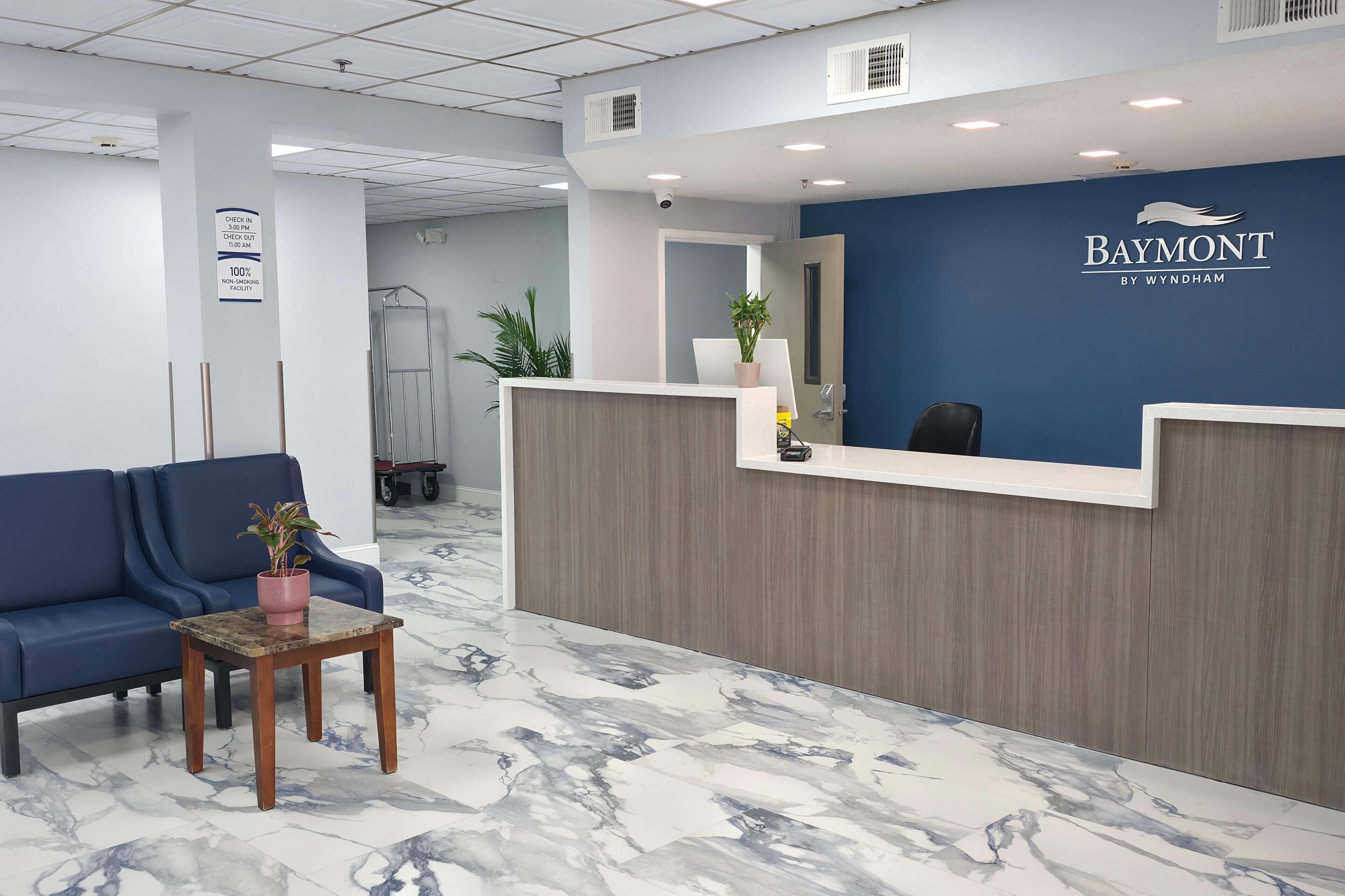 Ein Empfangsbereich mit blauer Wand und einem Schild mit der Aufschrift 'Baymont by Wyndham'. Ein blaues Sofa und ein kleiner Tisch mit einer Pflanze sind ebenfalls zu sehen.