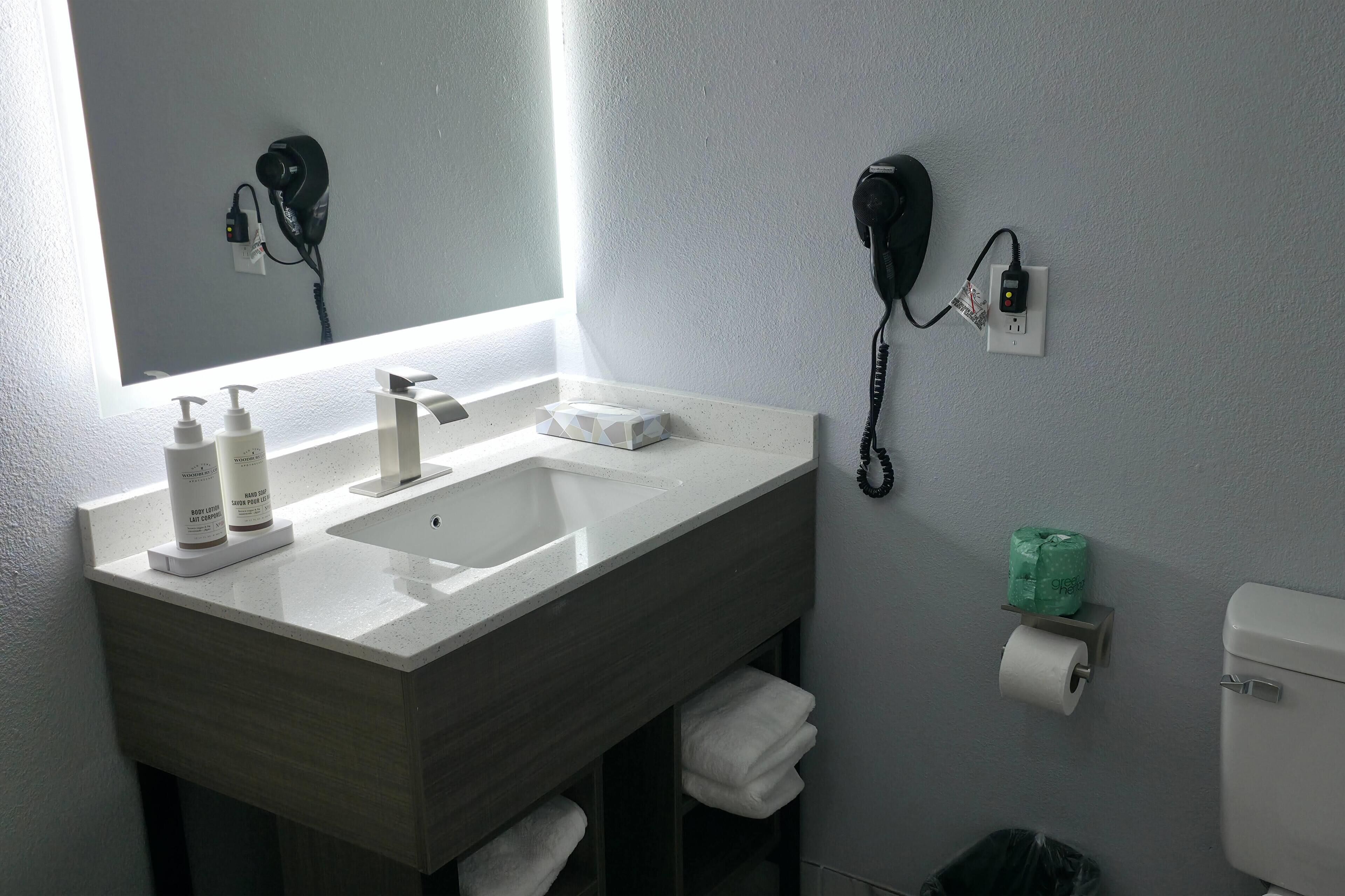 Un baño con lavamanos, dos secadores de pelo, un espejo, inodoro y un cubo de basura.