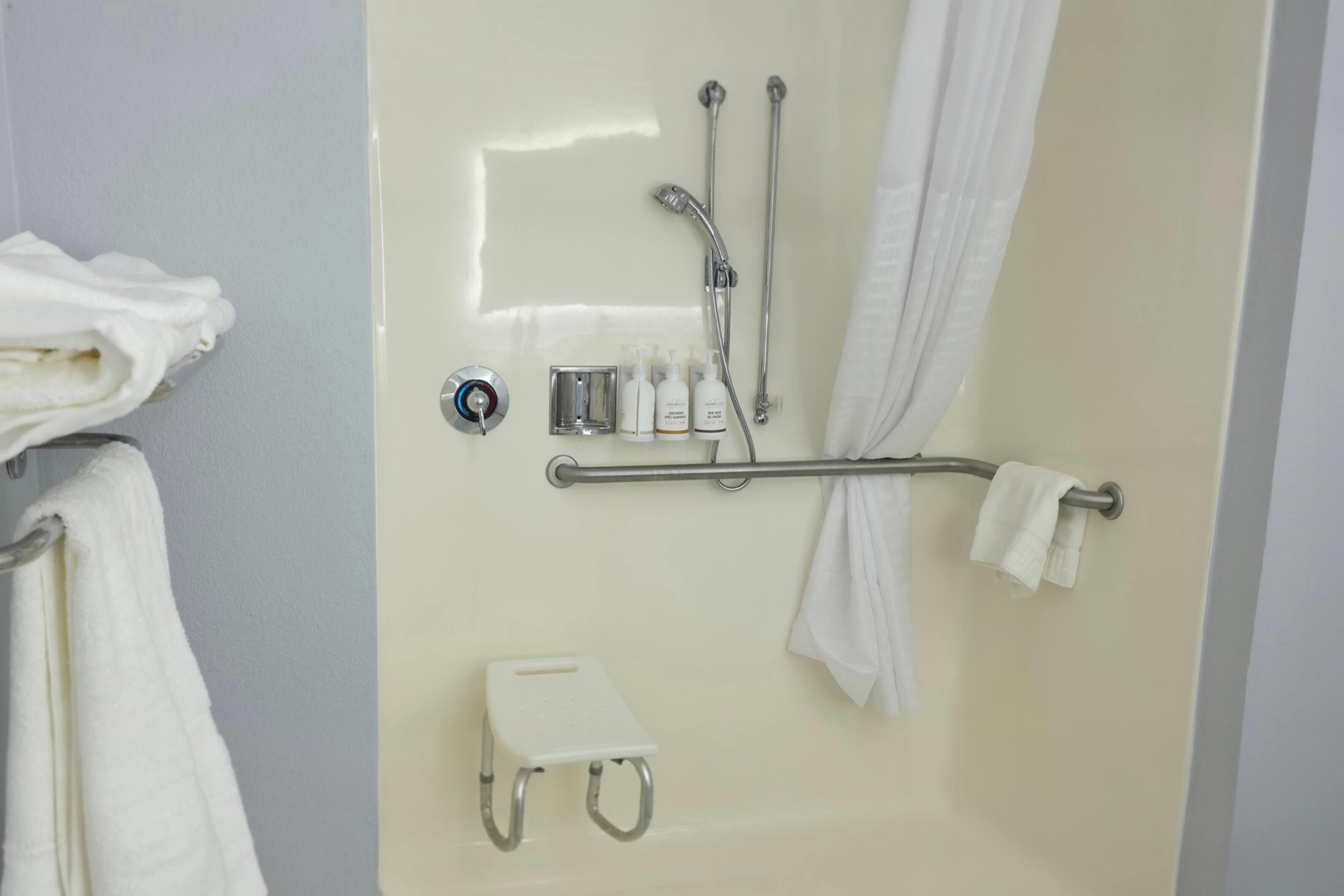 Baño con ducha, barras de apoyo, toallero y un pequeño taburete.
