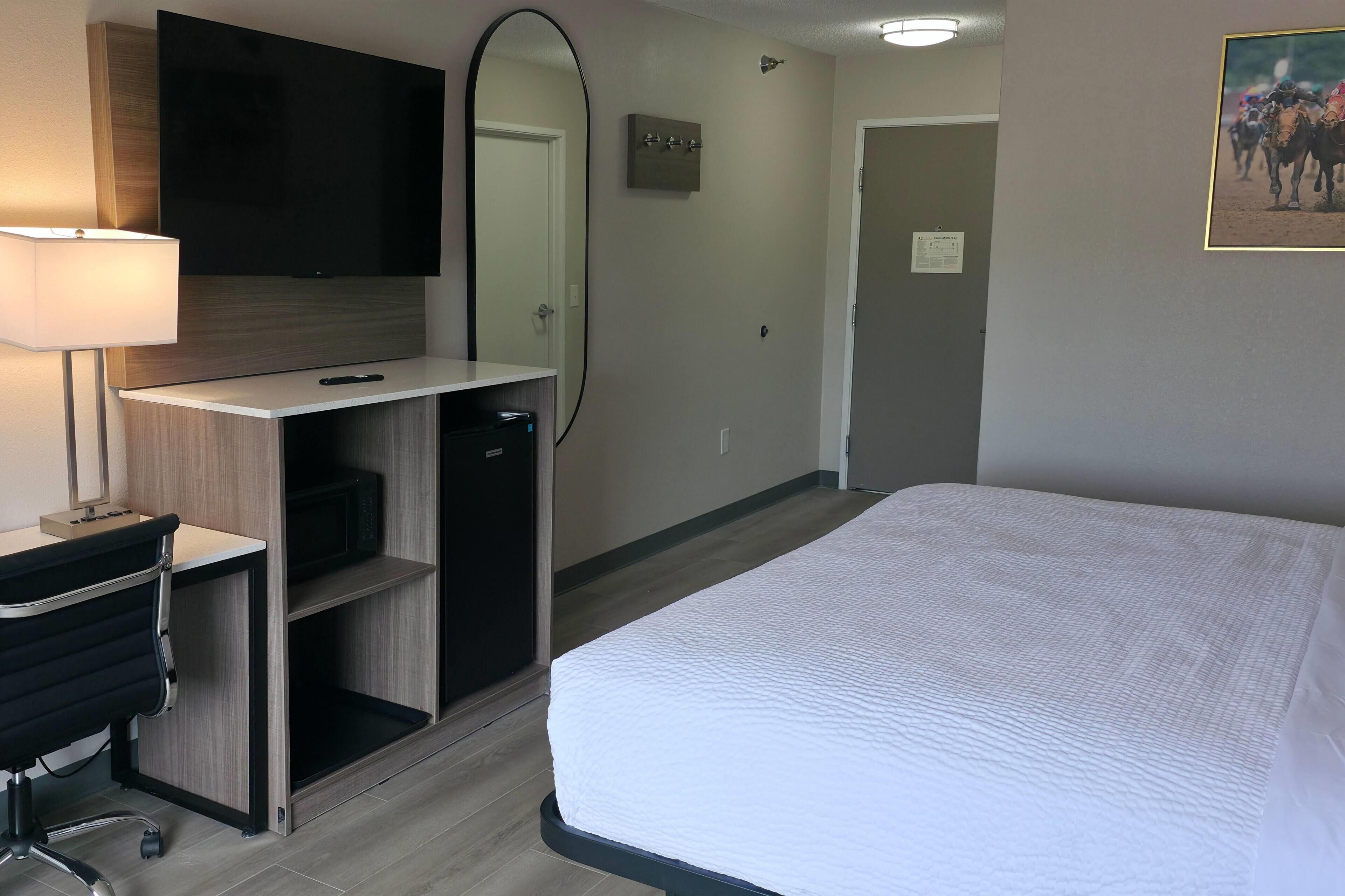 Quarto de hotel com uma cama, uma escrivaninha com luminária, um espelho e uma televisão.