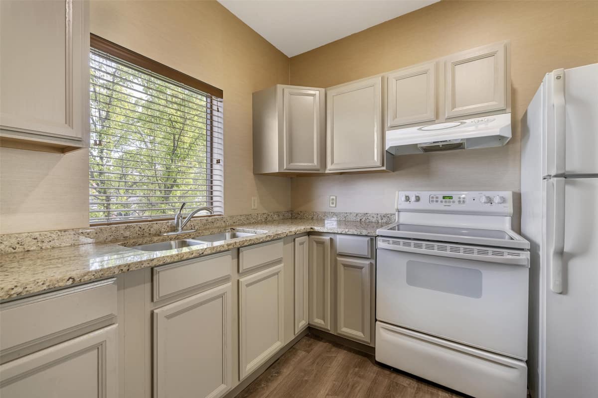 A kitchen with white cabinets, a granite countertop, a built-in oven, and a window with blinds.