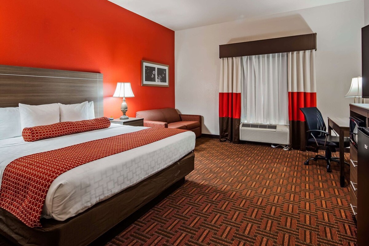 A hotel room with a large bed, a desk, a chair, a desk lamp, a picture frame, and a window with curtains.