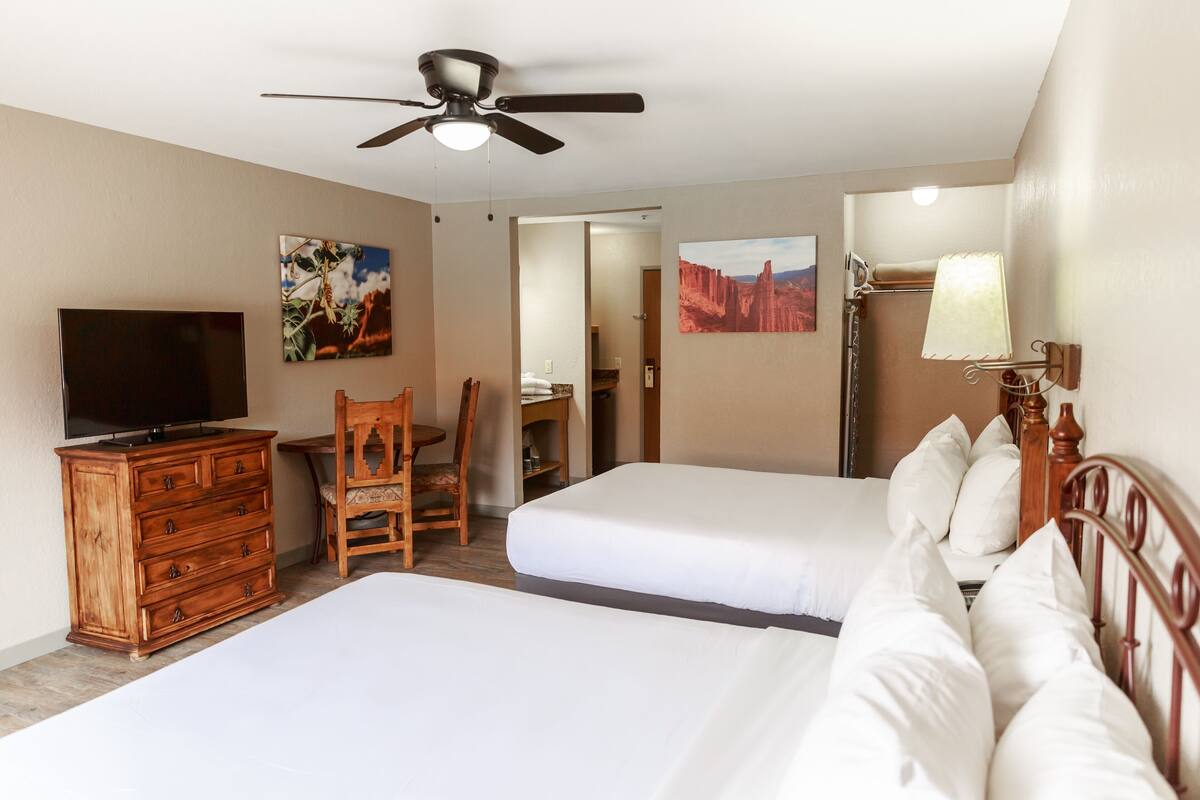 A hotel room with two beds, a wooden dresser, a television, a ceiling fan, and artwork on the walls.