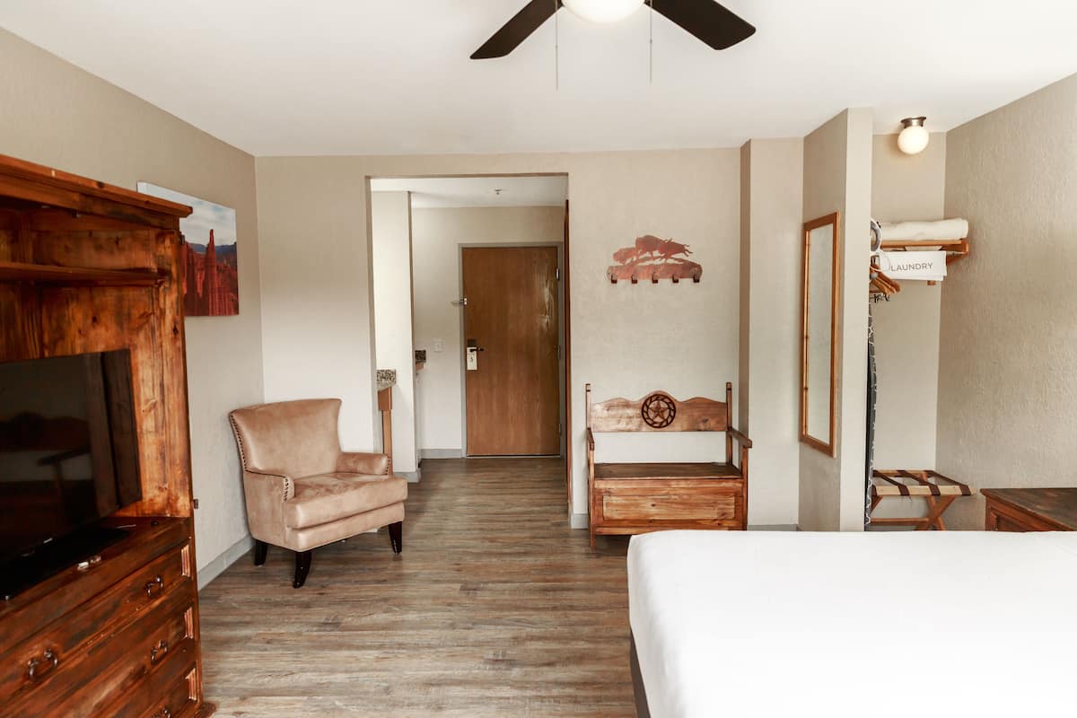 A hotel room with a wooden dresser, a leather armchair, a wooden bench, a bed, a ceiling fan, and a wall-mounted laundry rack.