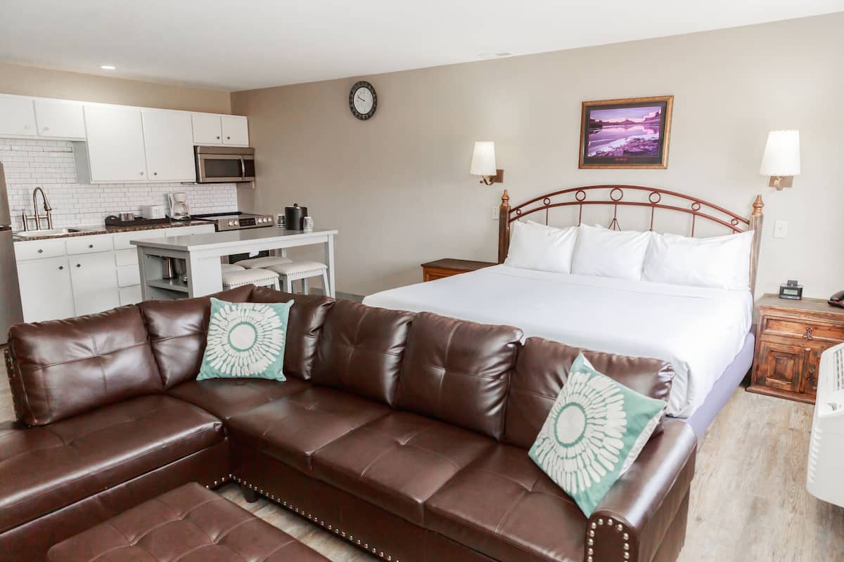 A modern hotel room with a brown leather sofa, a large bed with white linens, a kitchenette, and a painting on the wall.