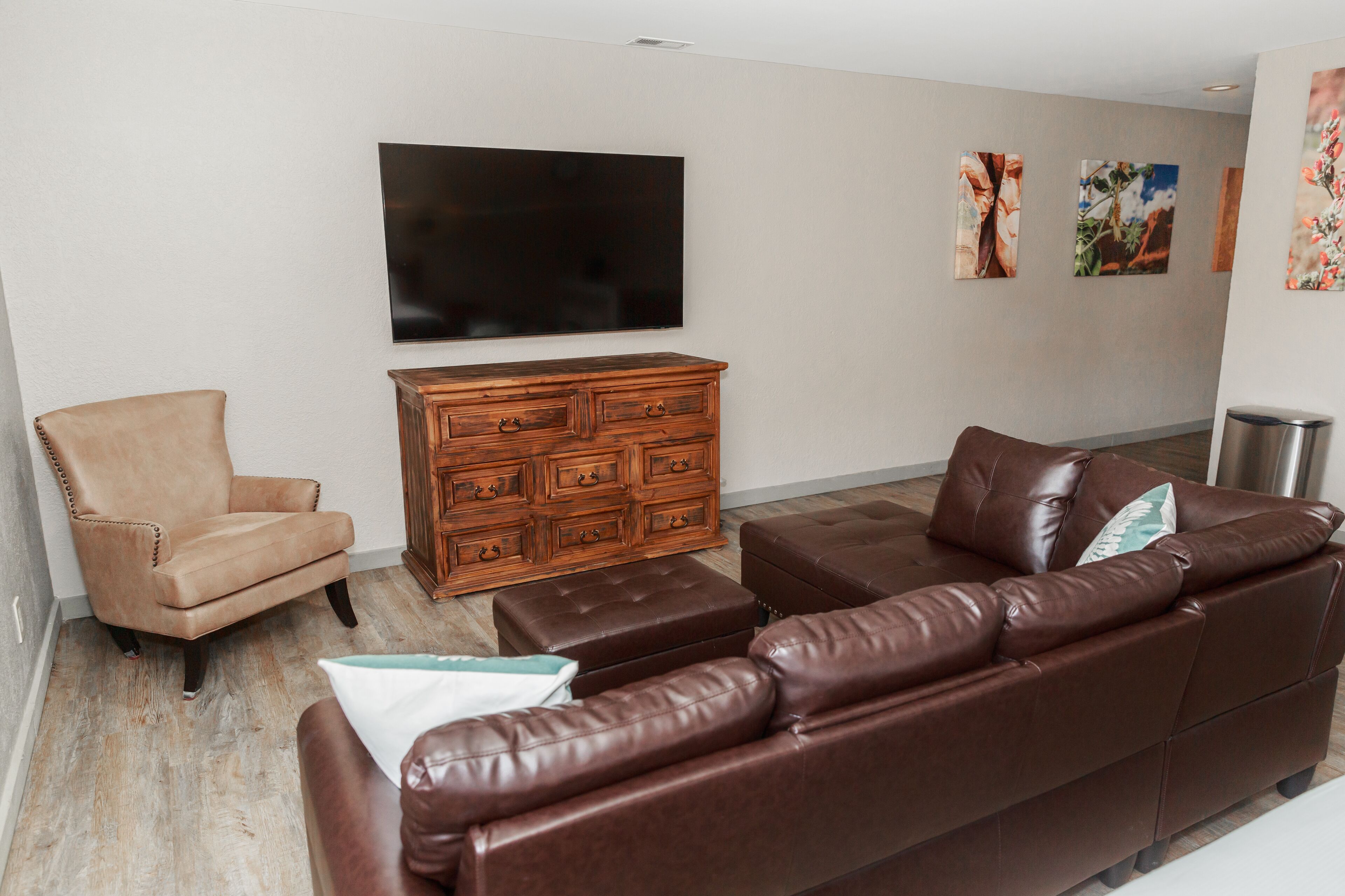 A living room with a brown leather sofa, a wooden cabinet, a beige armchair, and a flat-screen TV mounted on the wall.