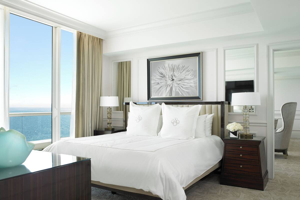A hotel room with a large bed, a nightstand, a desk, a chair, a TV, and a window with a view of the ocean.