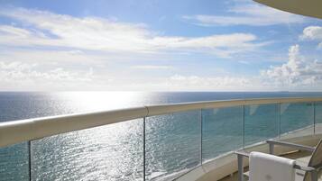Un balcón con vista al océano, barandilla de cristal y una silla.