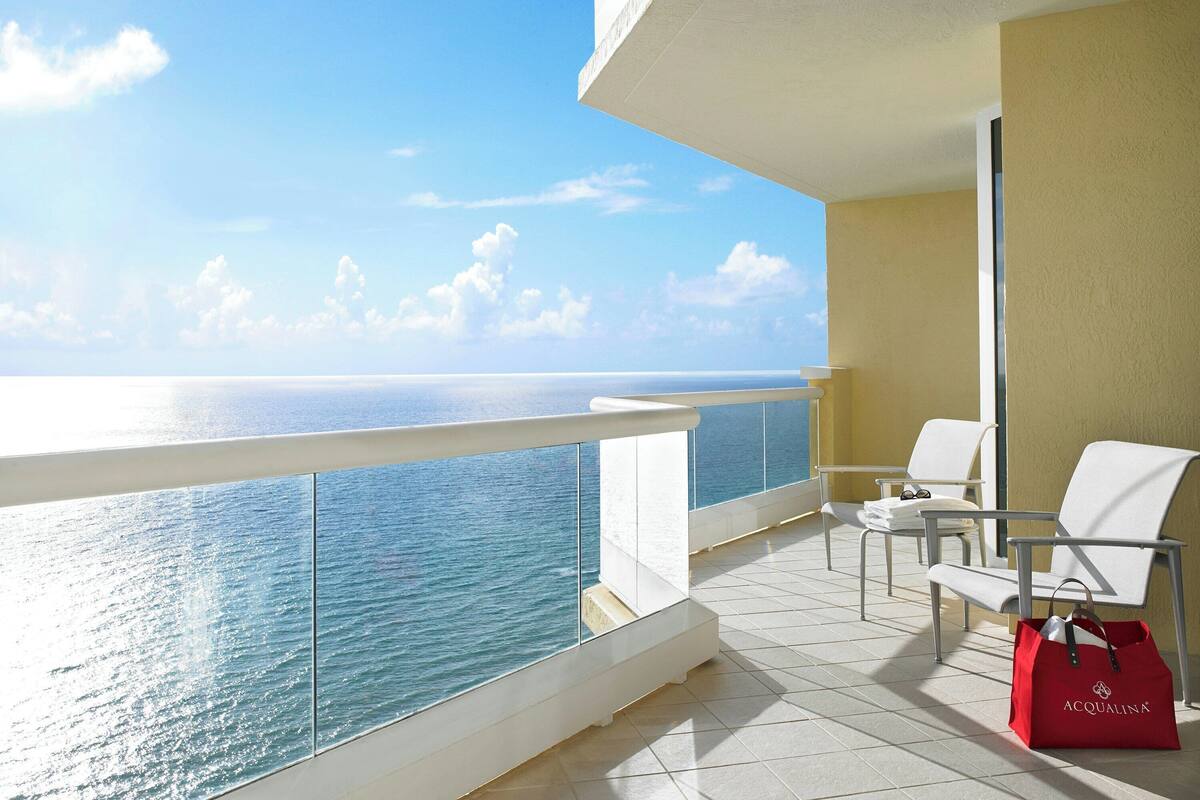 A balcony with a clear view of the ocean, white chairs, and a red bag.