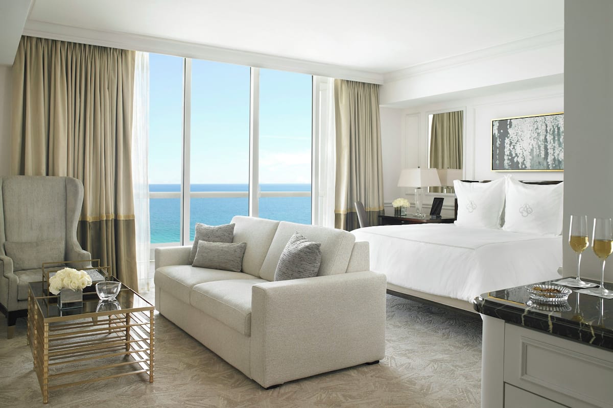 A hotel room with a large window, a sofa, a bed, a desk, and a minibar.
