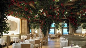 A restaurant with tables set for dining, a view of the sea through large windows, and a ceiling adorned with red flowers.