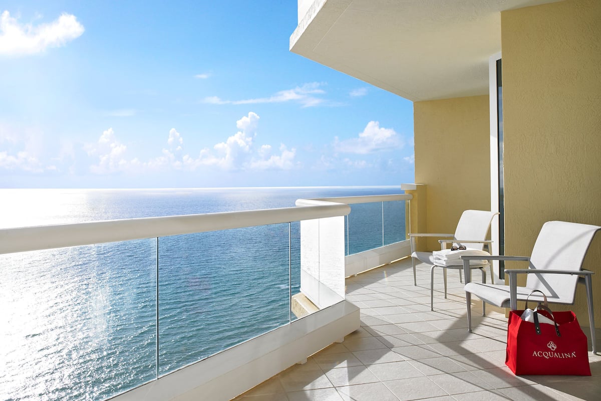 A balcony with a clear view of the ocean, white chairs, a red bag, and a yellow wall.