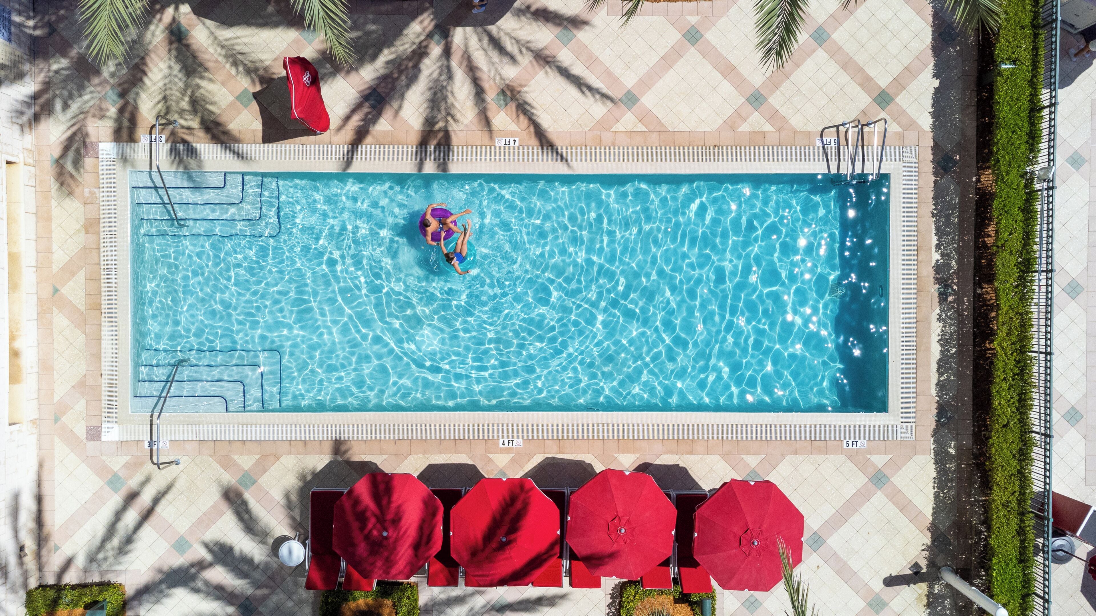 A rectangular swimming pool with clear blue water, surrounded by a tiled deck. There are red umbrellas and chairs arranged around the pool.