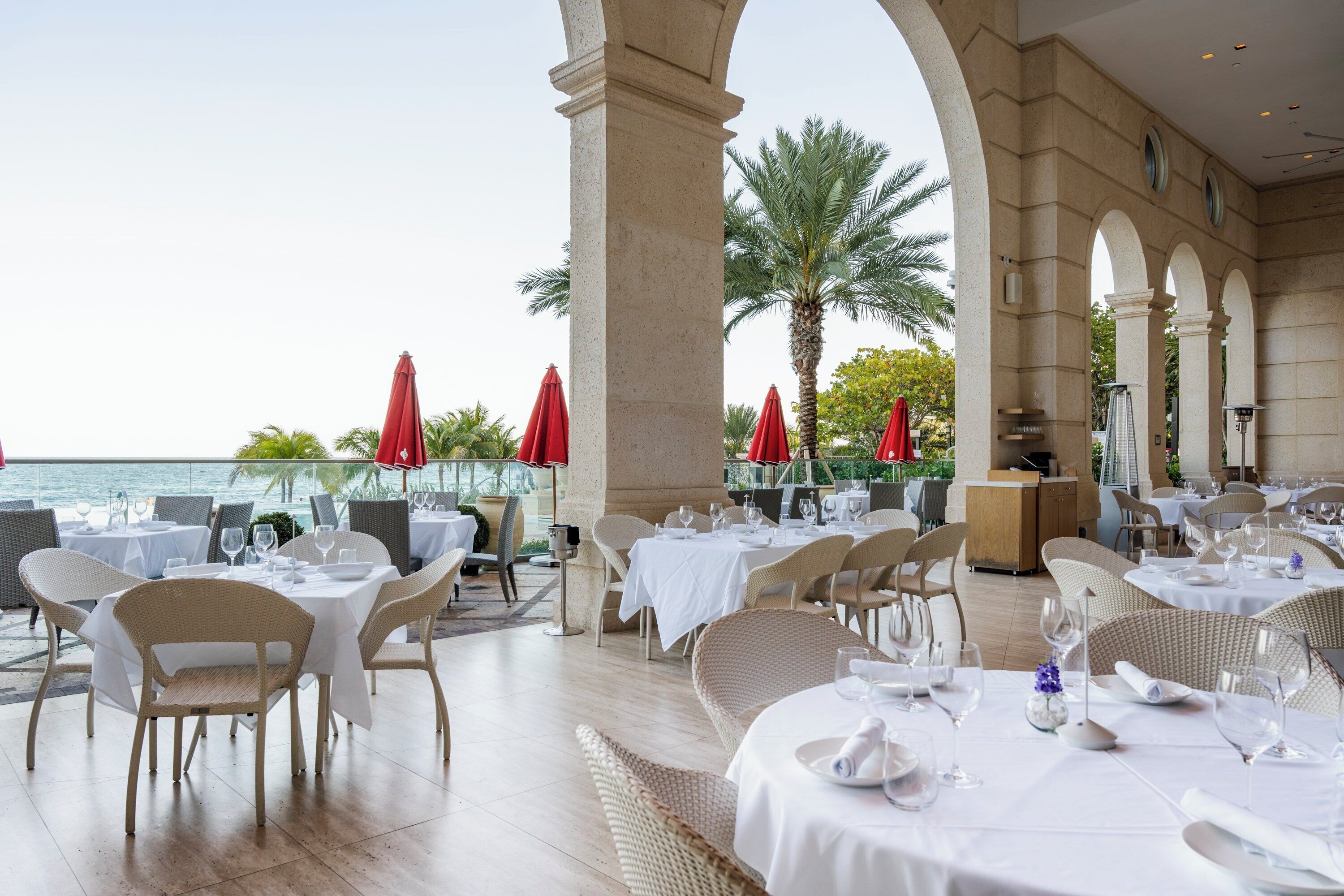 Un área de comedor al aire libre con mesas y sillas, palmeras y vistas al mar.