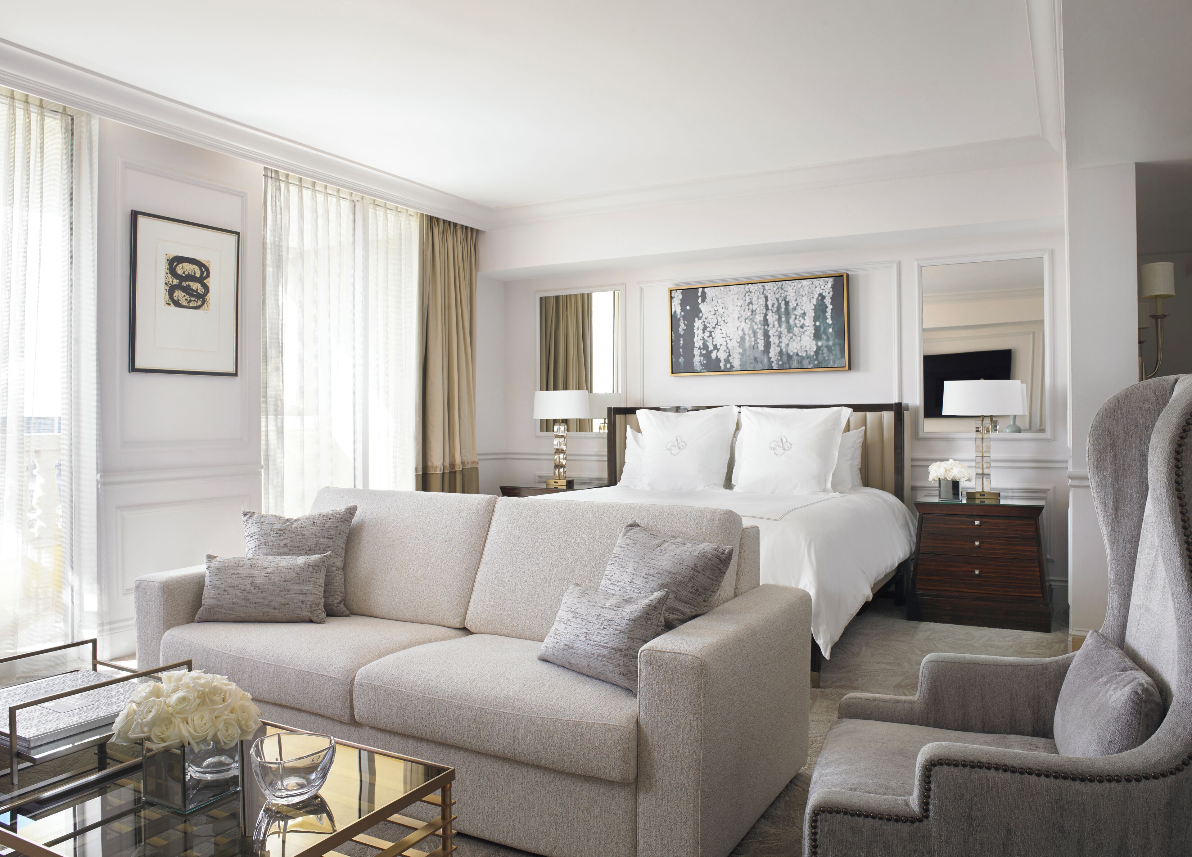Habitación de hotel con sofá, sillón y cama con ropa blanca. Hay un cuadro en la pared y un florero con flores sobre la mesa.