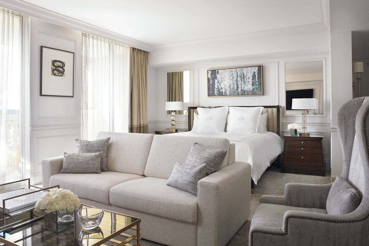 Habitación de hotel con sofá, sillón y cama con ropa blanca. Hay un cuadro en la pared y un florero con flores sobre la mesa.