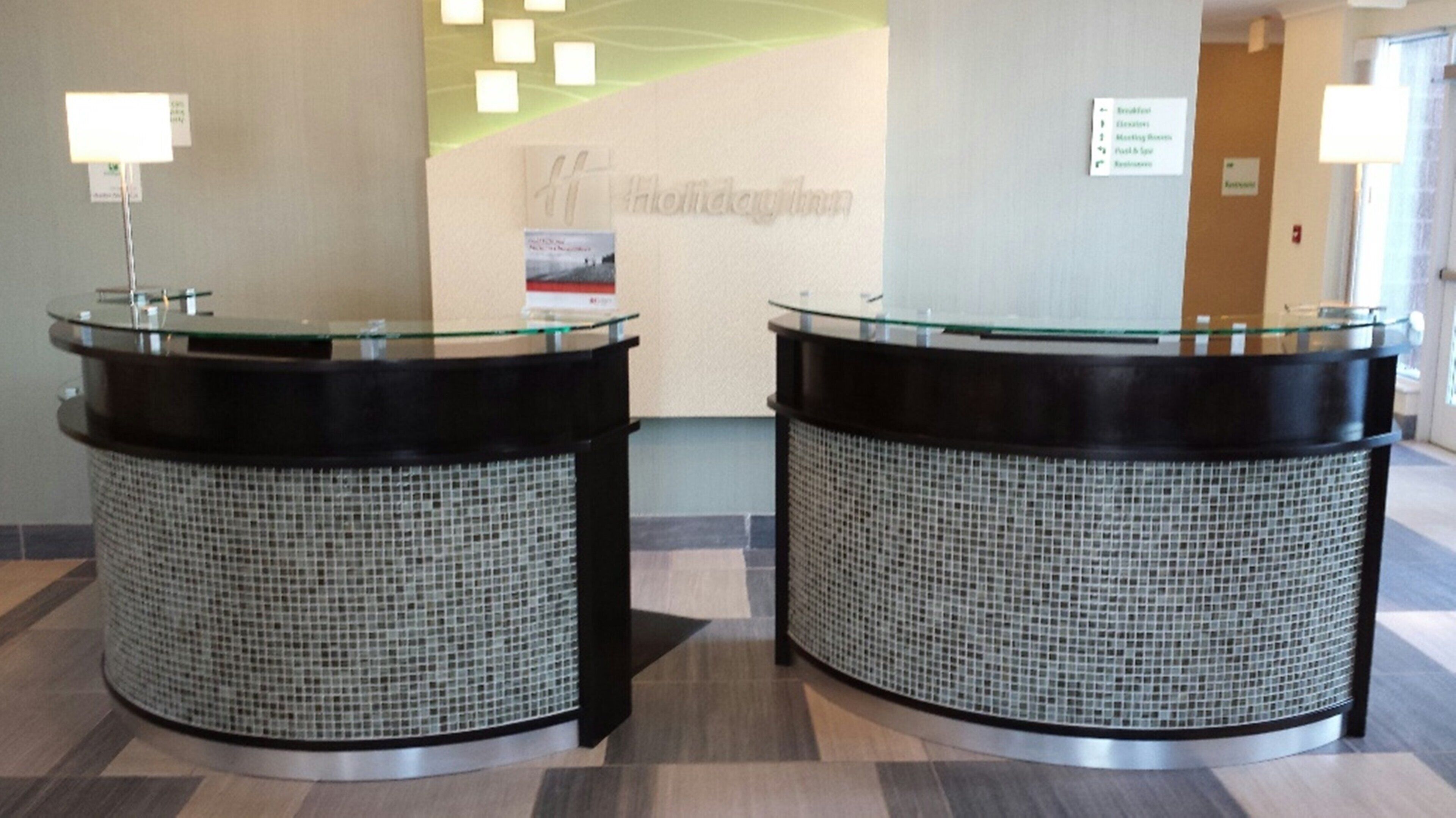 Two reception counters with glass tops and woven backs, a sign for 'Holiday Inn' in the background, and a sign listing hotel amenities.