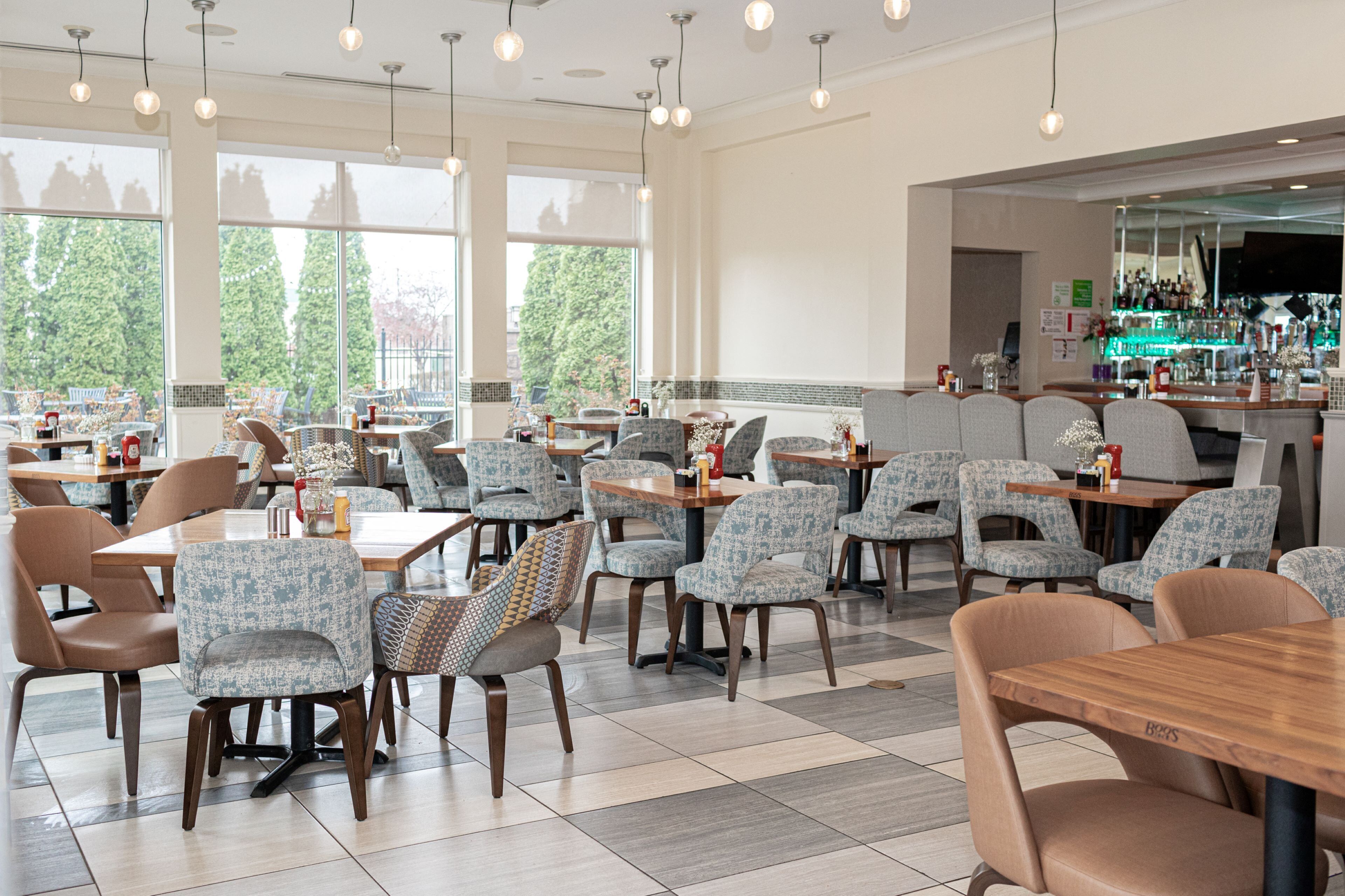 Un vaste espace dédié à la salle à manger, avec de nombreuses tables et chaises, de grandes fenêtres offrant une vue sur les arbres, et un coin bar doté d’un comptoir et d’étagères.