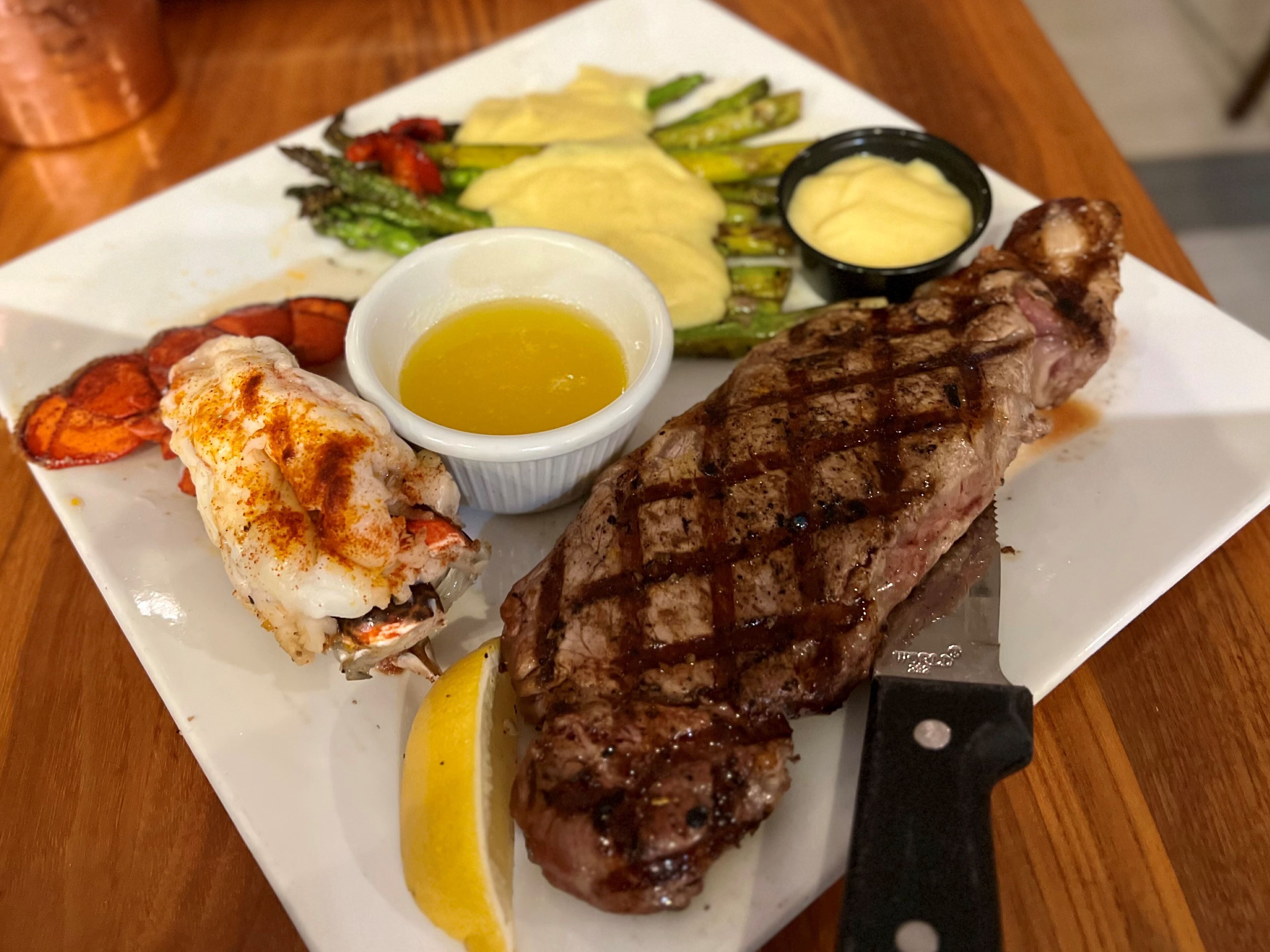 A steak with grill marks, a lobster tail, asparagus, and a lemon wedge on a white plate.