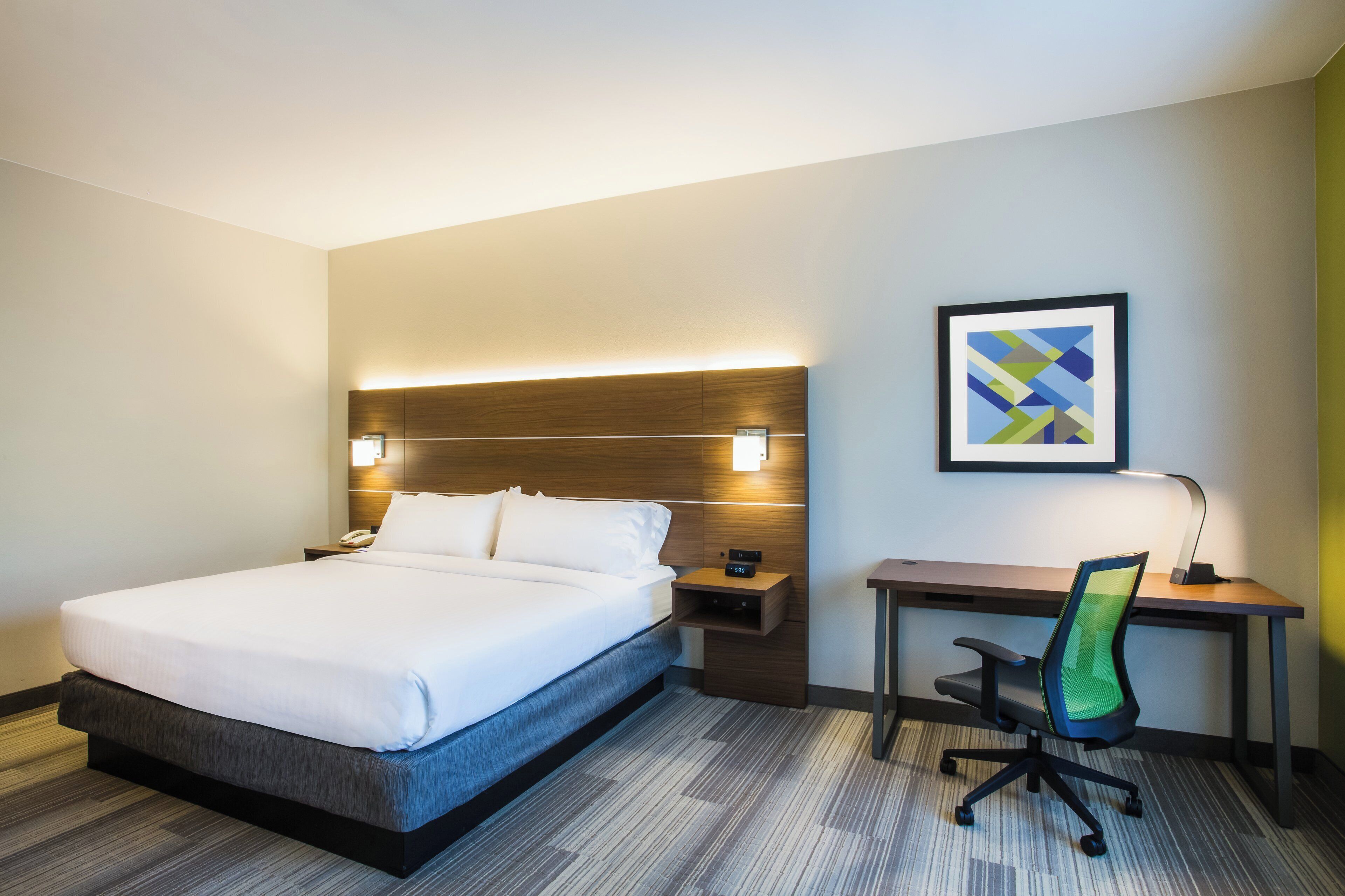 Una habitación de hotel con cama, mesita de noche, escritorio, silla y un cuadro en la pared.