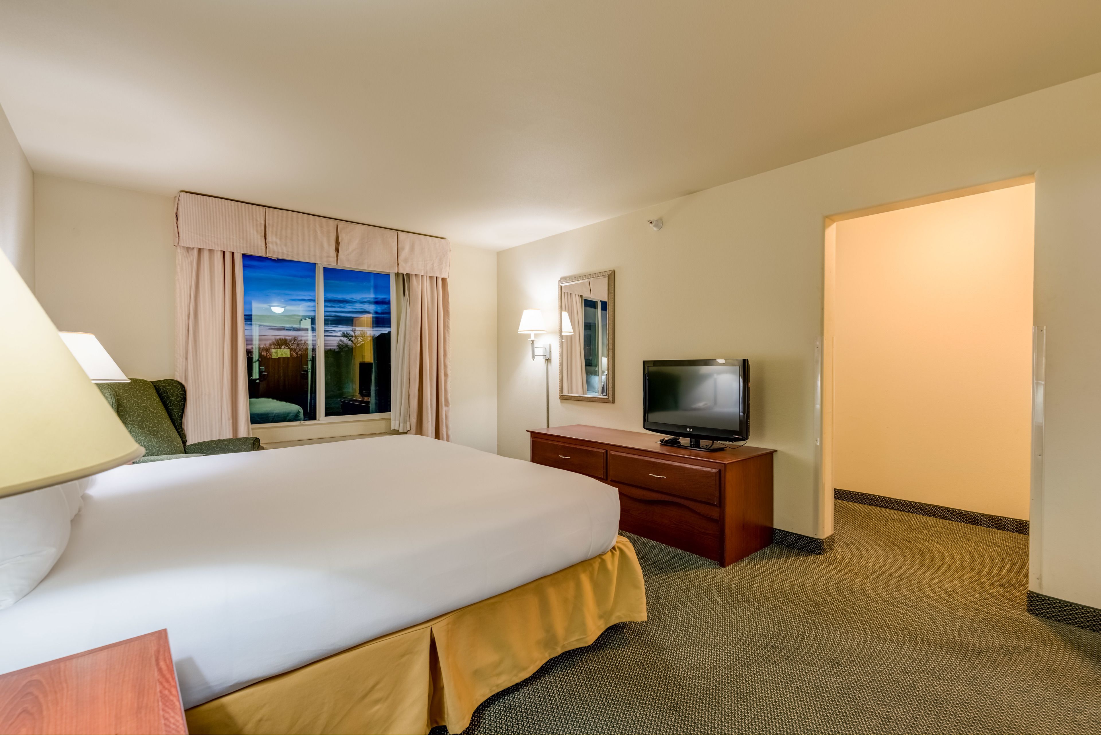 A hotel room with a large bed, a television on a wooden dresser, a window with curtains, and a carpeted floor.