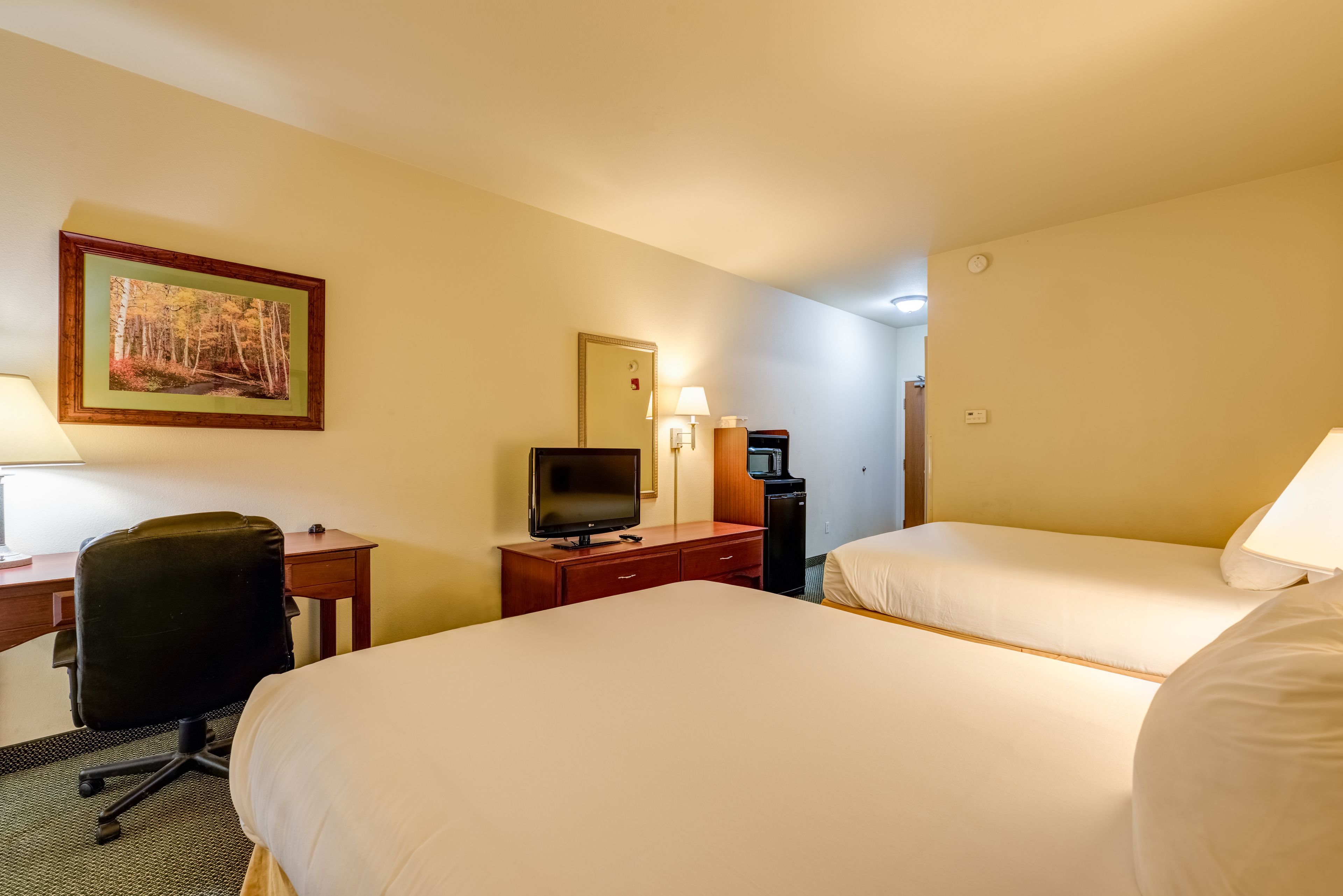 Habitación de hotel con dos camas, un escritorio con computadora, televisor, cafetera y un cuadro en la pared.